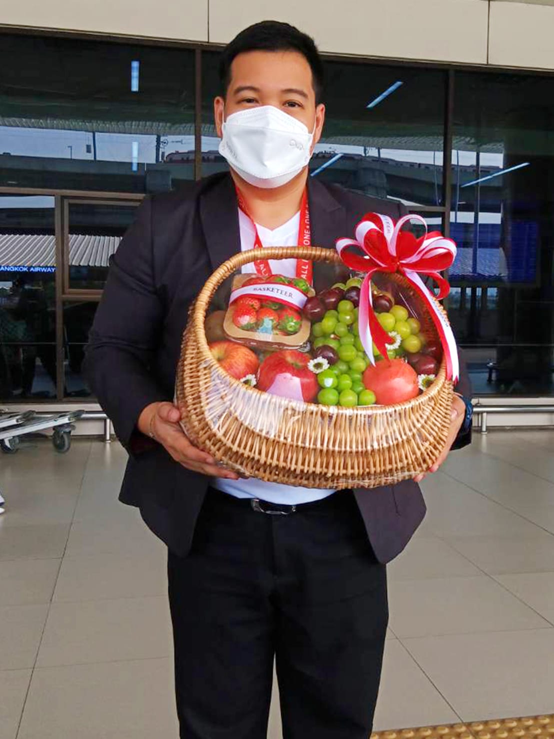 A person wearing a white mask, a white shirt, a black jacket, and a red lanyard around the neck is holding a wicker basket filled with an assortment of fruit, including apples and grapes. The mixed fruit basket is decorated with a red and white ribbon.