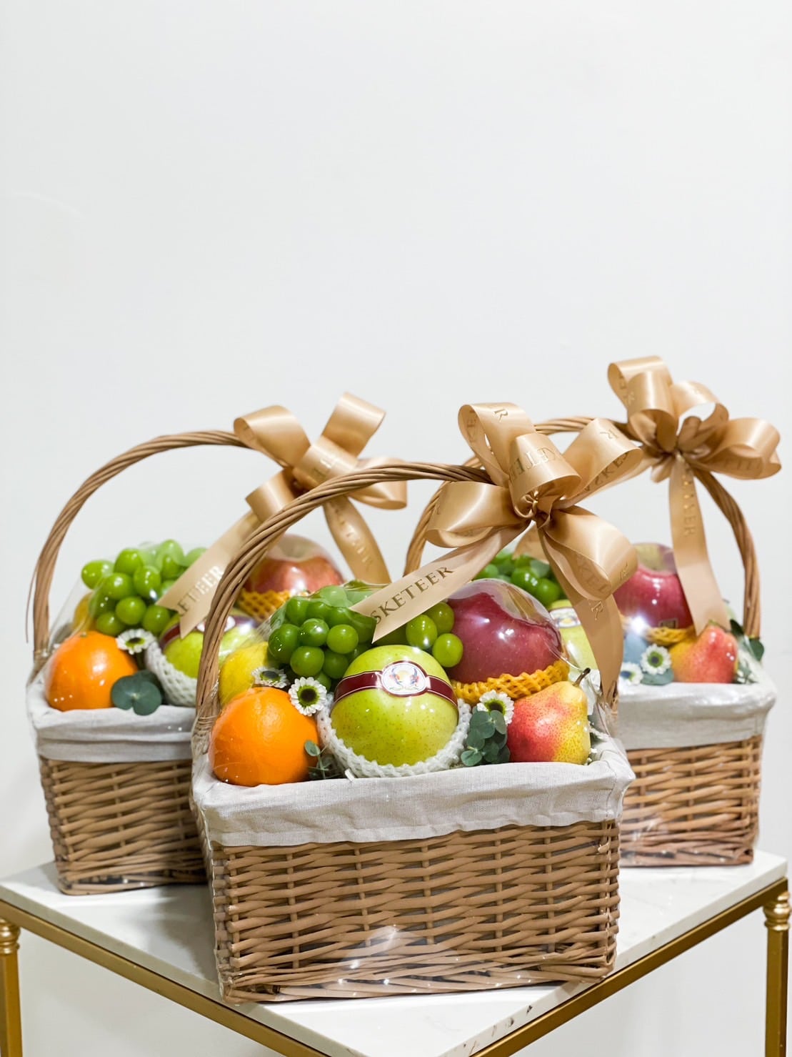 Three wicker baskets brimming with a lavish abundance of fresh fruits, including apples, grapes, oranges, and melons, each adorned with a large decorative gold ribbon tied into a bow. Placed atop a white surface, the baskets are lined with cloth and accented by a few small flowers.