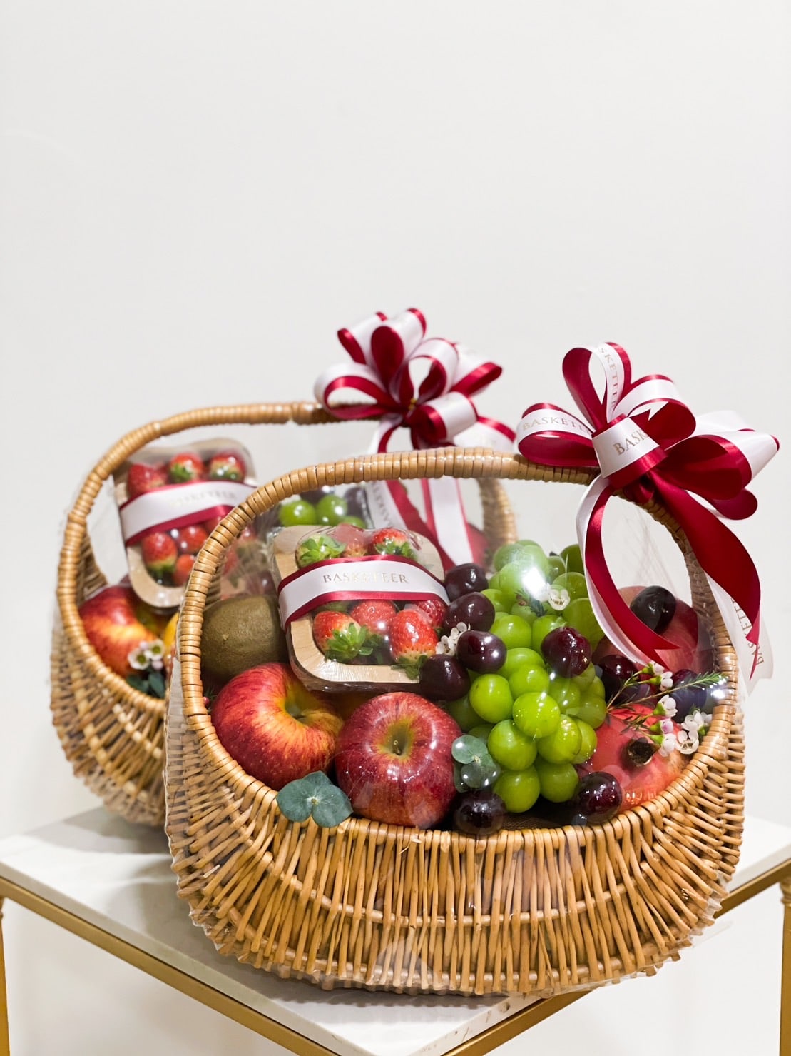 Two wicker baskets filled with an assortment of fresh fruits, including apples, grapes, kiwis, and strawberries. Each mixed fruit basket is wrapped in clear plastic and topped with a decorative red and white ribbon bow. They are placed side by side on a white surface.