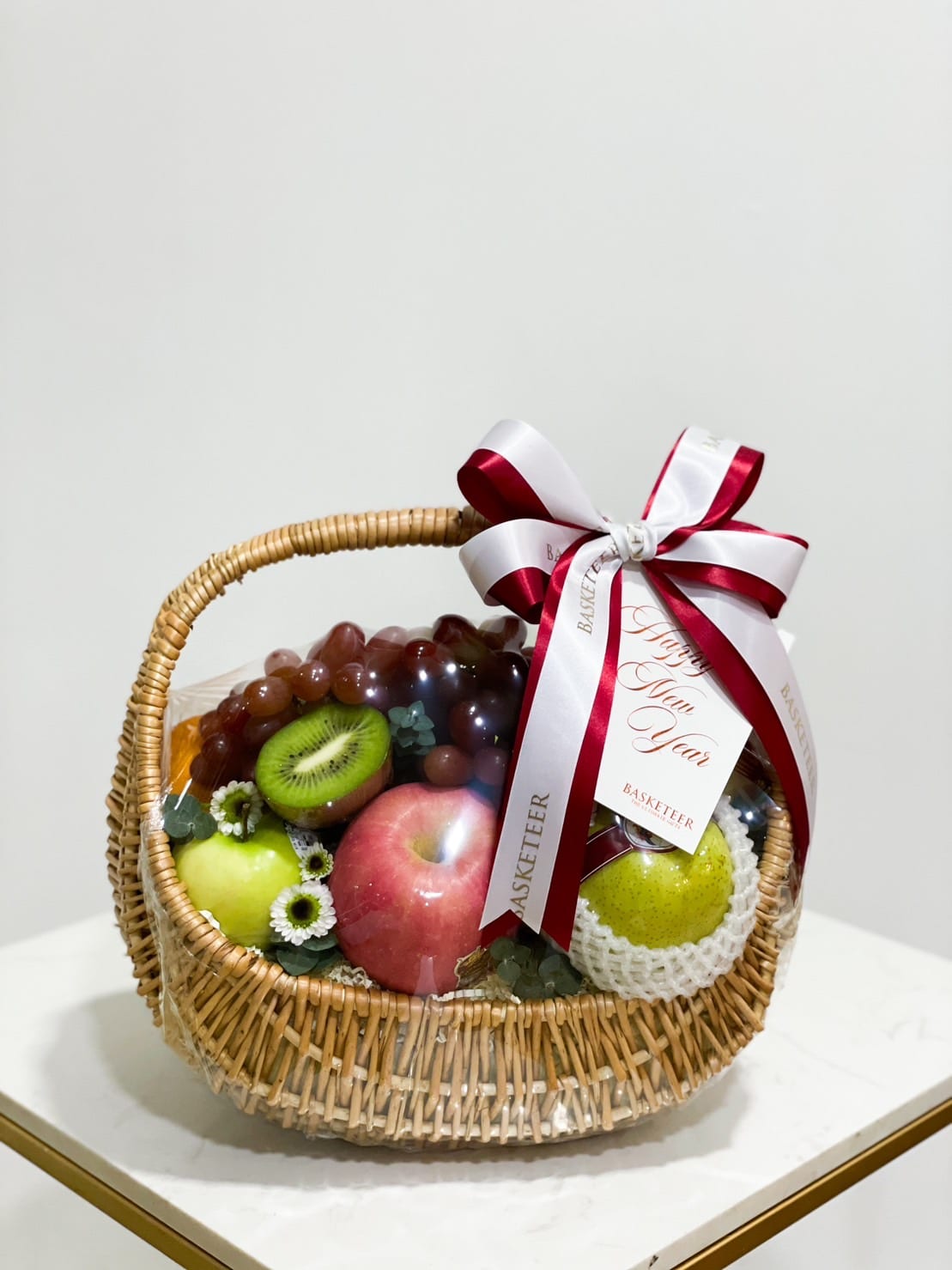 A wicker Fruit Ensemble Gift Basket filled with assorted fruits including apples, grapes, kiwis, and an orange, adorned with white and red ribbons. The ribbons have messages and branding, wishing a 