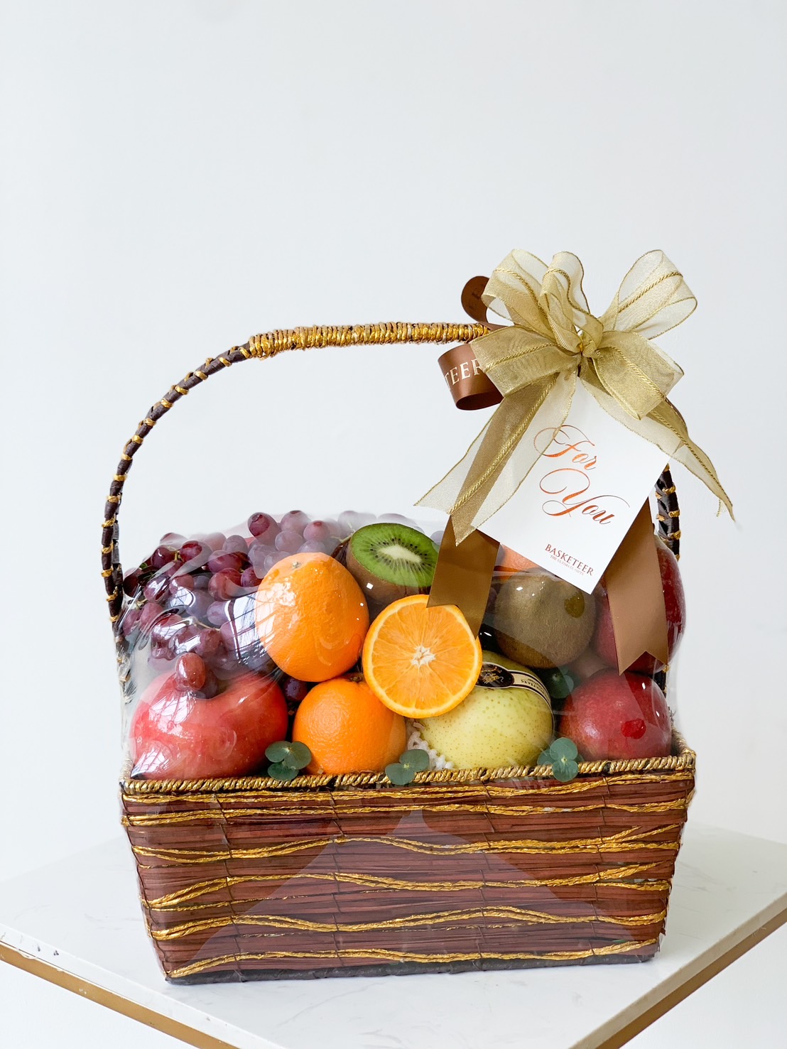 A woven basket from the Fruitopia Selections Gift Basket collection, filled with fresh apples, oranges, kiwis, and grapes. Wrapped in clear plastic and adorned with a gold and brown ribbon with a 
