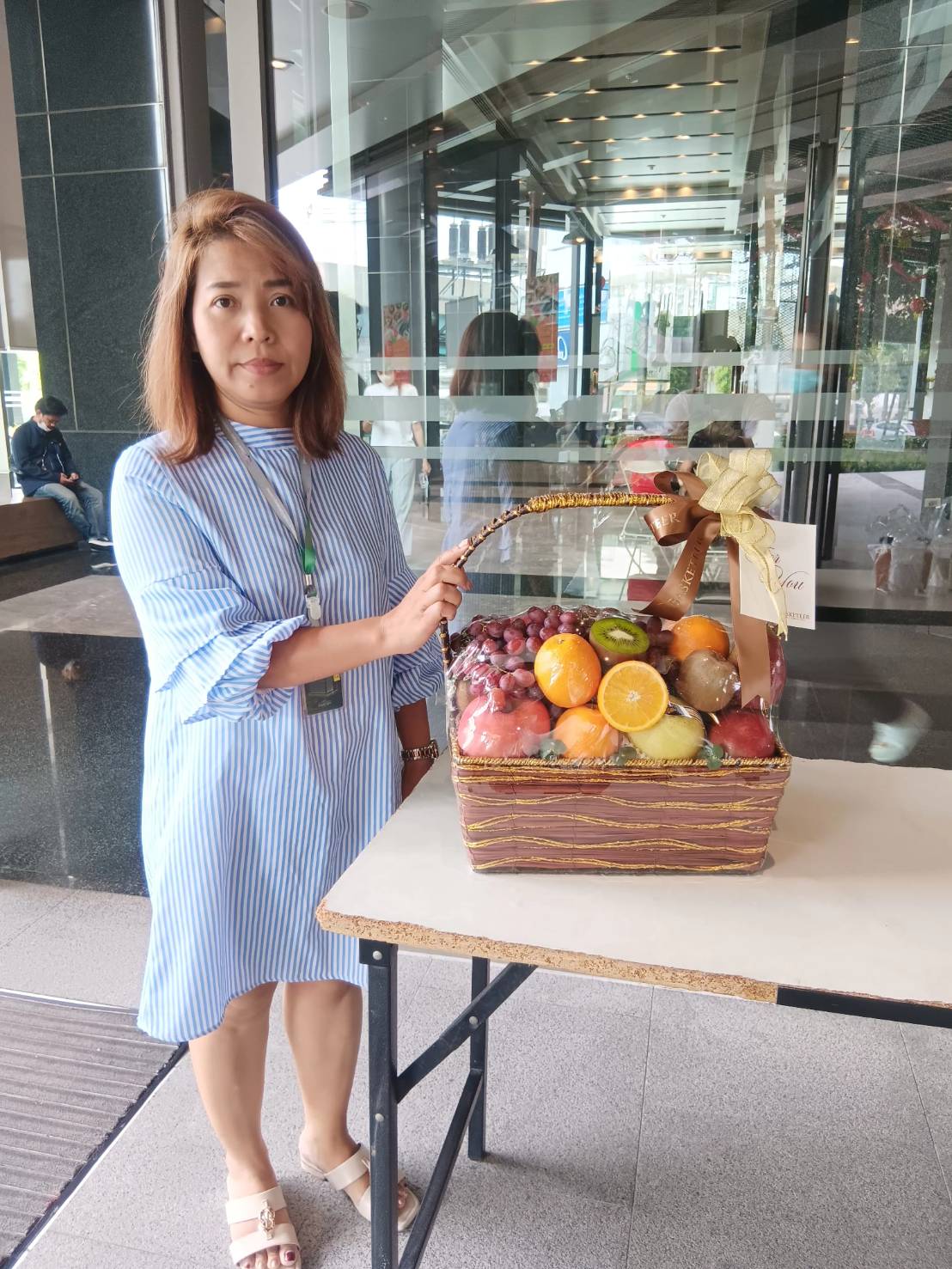 A woman in a blue striped dress stands next to a table holding a Fruitopia Selections Gift Basket, brimming with an assortment of fruits like grapes, oranges, and apples. She has shoulder-length brown hair, and the background reveals the modern lobby of a building with glass windows and doors.