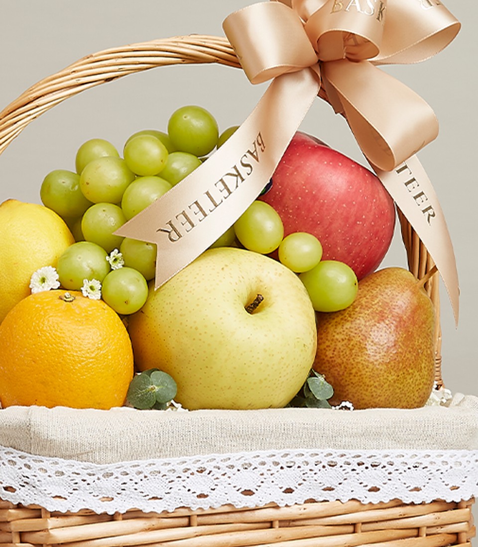 A rattan gift basket Filled with a variety of fresh fruits such as green grapes, red apples, oranges, and many more. Decorated with a bow. Beautiful gold ribbon with the word “Basketeer” on it.