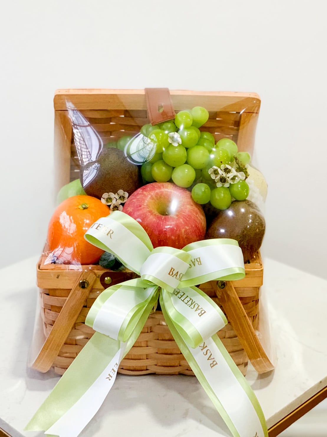 The Gourmet Paradise Fruit Basket is a wicker gift basket filled with assorted fruits like green grapes, a red apple, an orange, and kiwis. Wrapped in clear plastic and tied with a white and green ribbon bow, this basket is beautifully presented on a white surface against a light background.