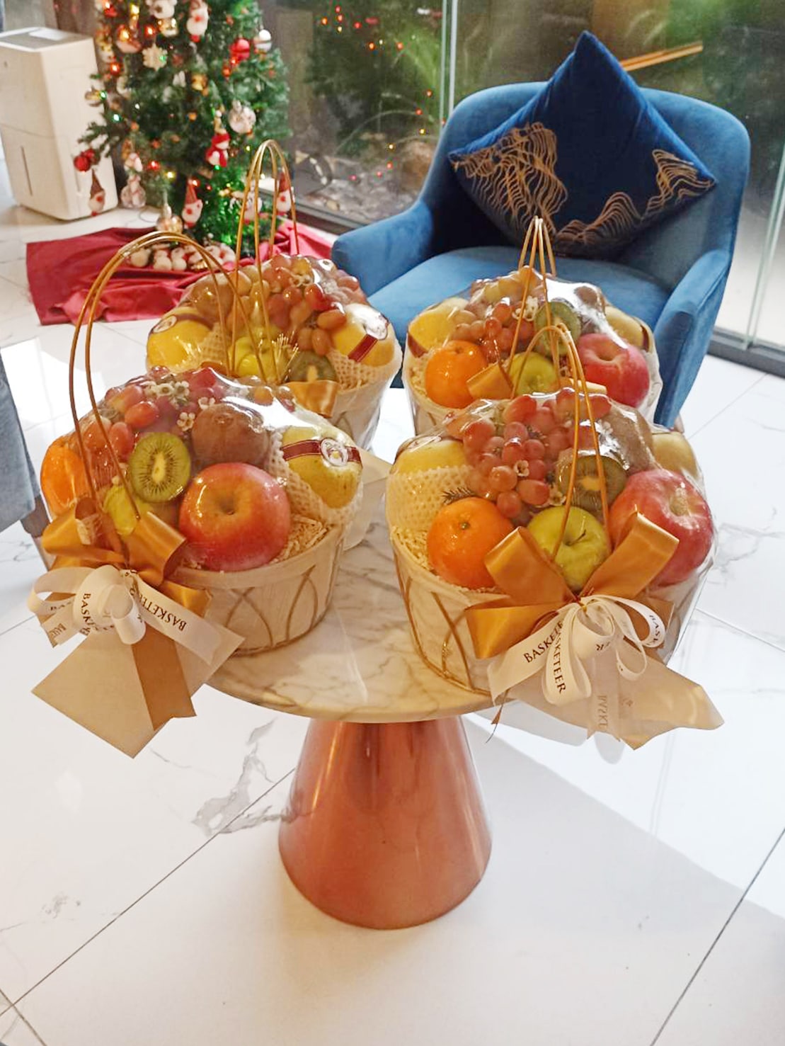 Four baskets of assorted fresh fruits—apples, oranges, grapes, and kiwis—are neatly arranged on a round marble table with a rose gold base. Each Fruit Basket Deluxe Treat is wrapped in cellophane and adorned with a golden ribbon. A Christmas tree is visible in the background.