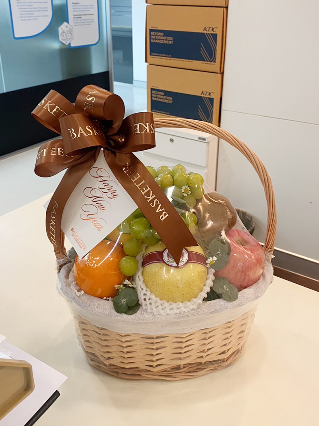 A wicker basket filled with various fruits including apples, oranges, and grapes, wrapped in clear cellophane. The basket is adorned with a brown ribbon that reads 
