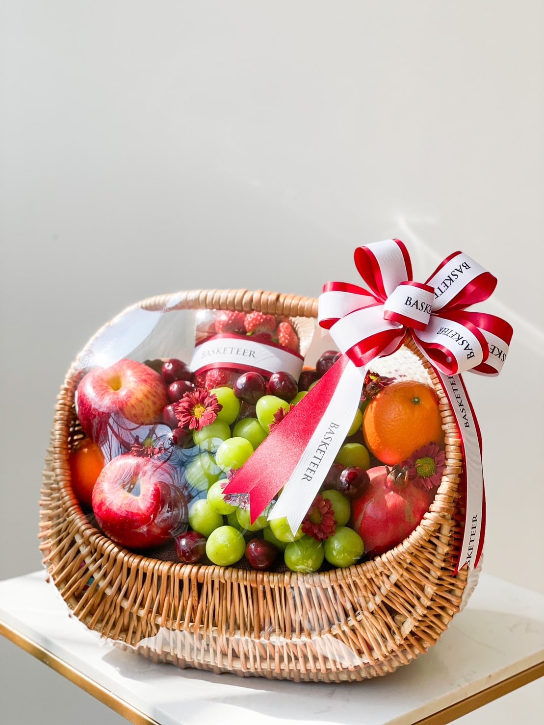 A wicker basket filled with assorted fruits including apples, grapes, and oranges, wrapped in clear cellophane. The mixed fruit basket is adorned with a red and white ribbon bow with 