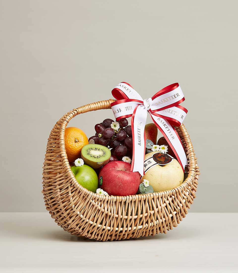 A Brown Rattan Gift Basket Filled with a variety of fresh fruits such as green avocados. Yellow pears, red grapes, and so much more. It is decorated with a red and white ribbon bow with the word 