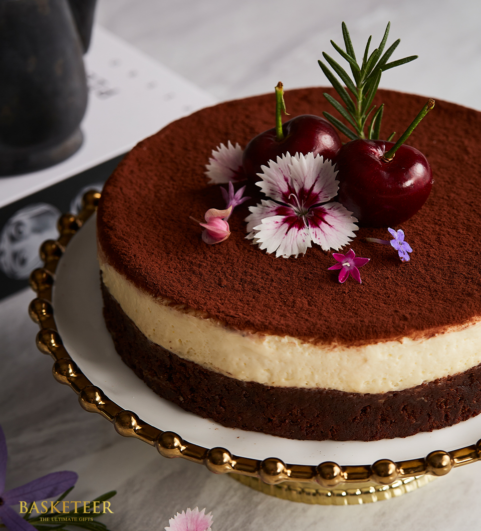 A decadent chocolate and vanilla layered cake, crowned with a light dusting of premium cocoa powder, garnished with fresh cherries, elegant pink and white flowers, and a fragrant sprig of rosemary is showcased on an exquisite decorative cake stand. The background features a beautifully designed booklet accompanied by an artful flower arrangement. In the scene, the 