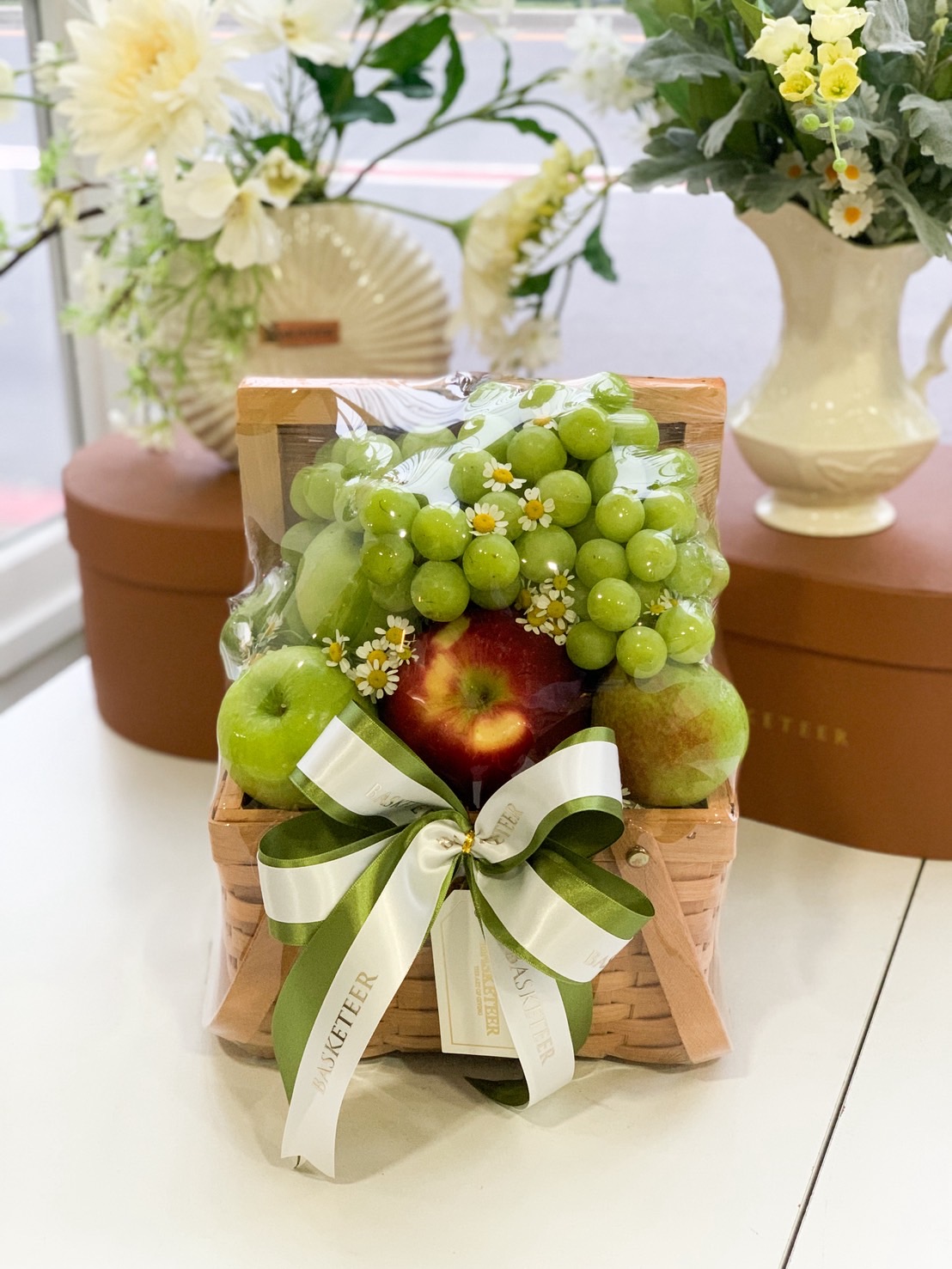 Premium fruit basket featuring green grapes, green and red apples, decorated with chamomile flowers and a green satin ribbon