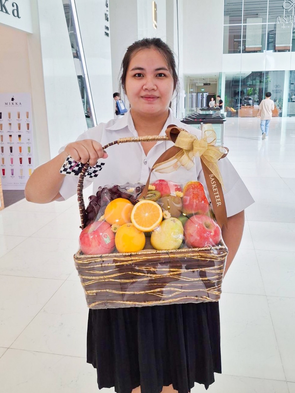 A woman in a white shirt and black skirt holds a Fruitopia Selections Gift Basket, filled with assorted fruits like apples, oranges, grapes, and lemons. The basket is adorned with a gold ribbon and bow. She stands in a bright, modern indoor space with people and displays in the background.
