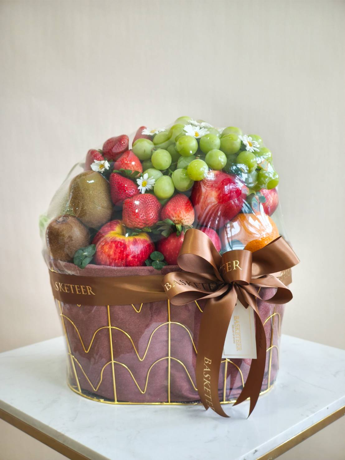 Luxury fruit basket with green grapes, strawberries, apples, kiwis, and chamomile flowers in a gold-wire frame with brown wrapping and bow
