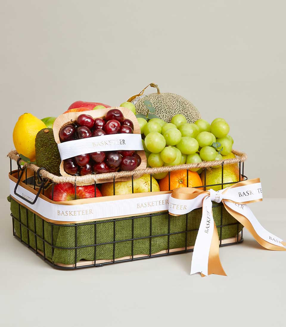 A sophisticated wire basket adorned with an elegant green fabric cradles a selection of orchard-fresh fruits, including luscious green grapes, vibrant cherries, crisp apples, juicy peaches, a perfectly ripe cantaloupe, and a zesty lime. The scene is enhanced by the white and gold ribbons that gracefully encircle the basket, proudly displaying the distinguished 