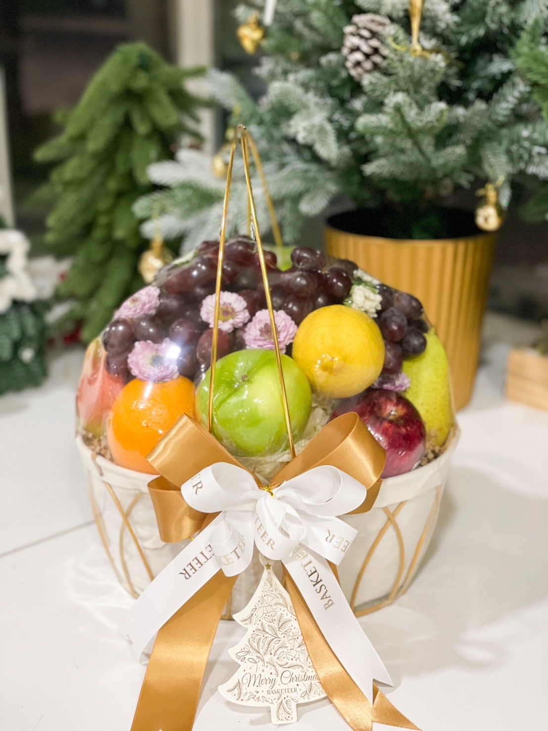 A beautifully arranged fruit basket featuring fresh apples, grapes, oranges, pears, and kiwis, elegantly presented in a golden wire basket lined with white fabric, adorned with a white and gold ribbon from Basketeer, perfect for gifting.