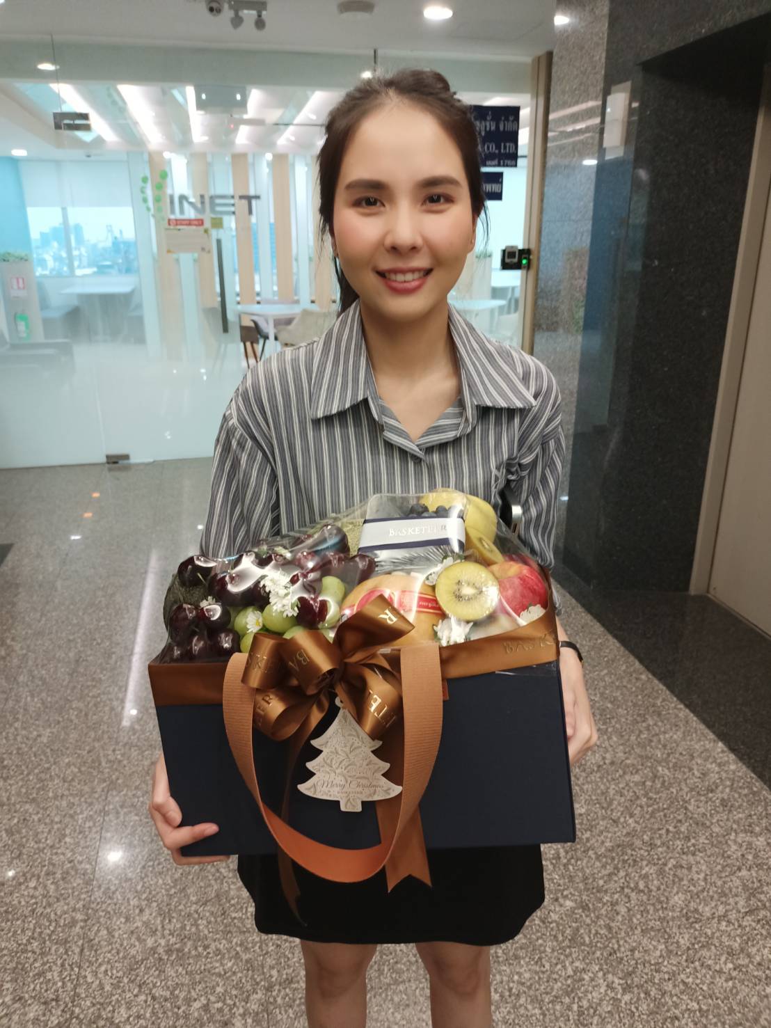 A smiling woman holding a premium fruit gift box by Basketeer, featuring fresh grapes, kiwis, and apples, elegantly wrapped with a brown ribbon and a festive Christmas tree tag, standing in a modern office setting.