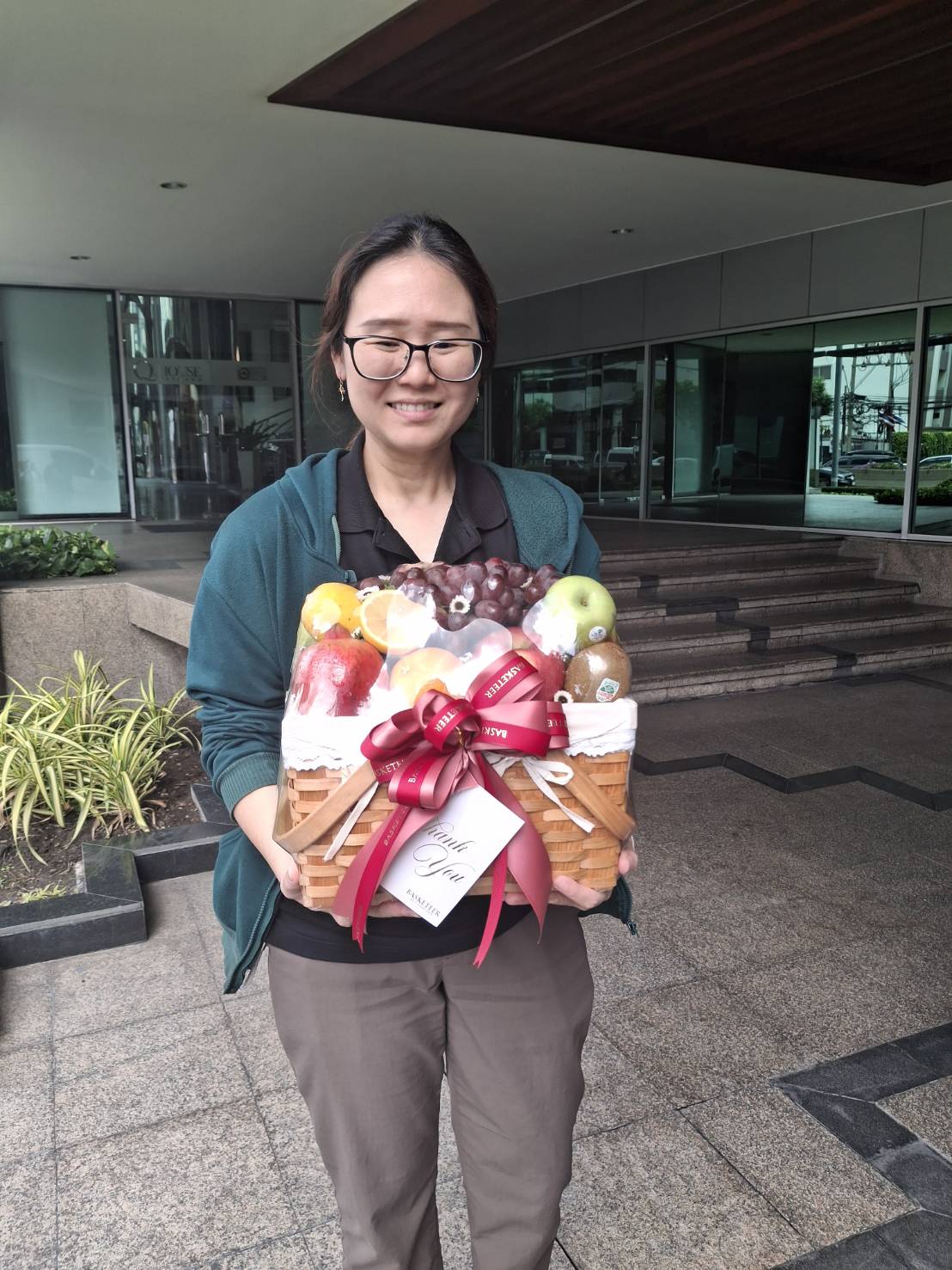 A person wearing glasses and a dark sweater smiles while holding a large Gala Luxury Fruit Basket, wrapped in clear plastic and adorned with a red ribbon. They stand outside a building near some greenery and steps.
