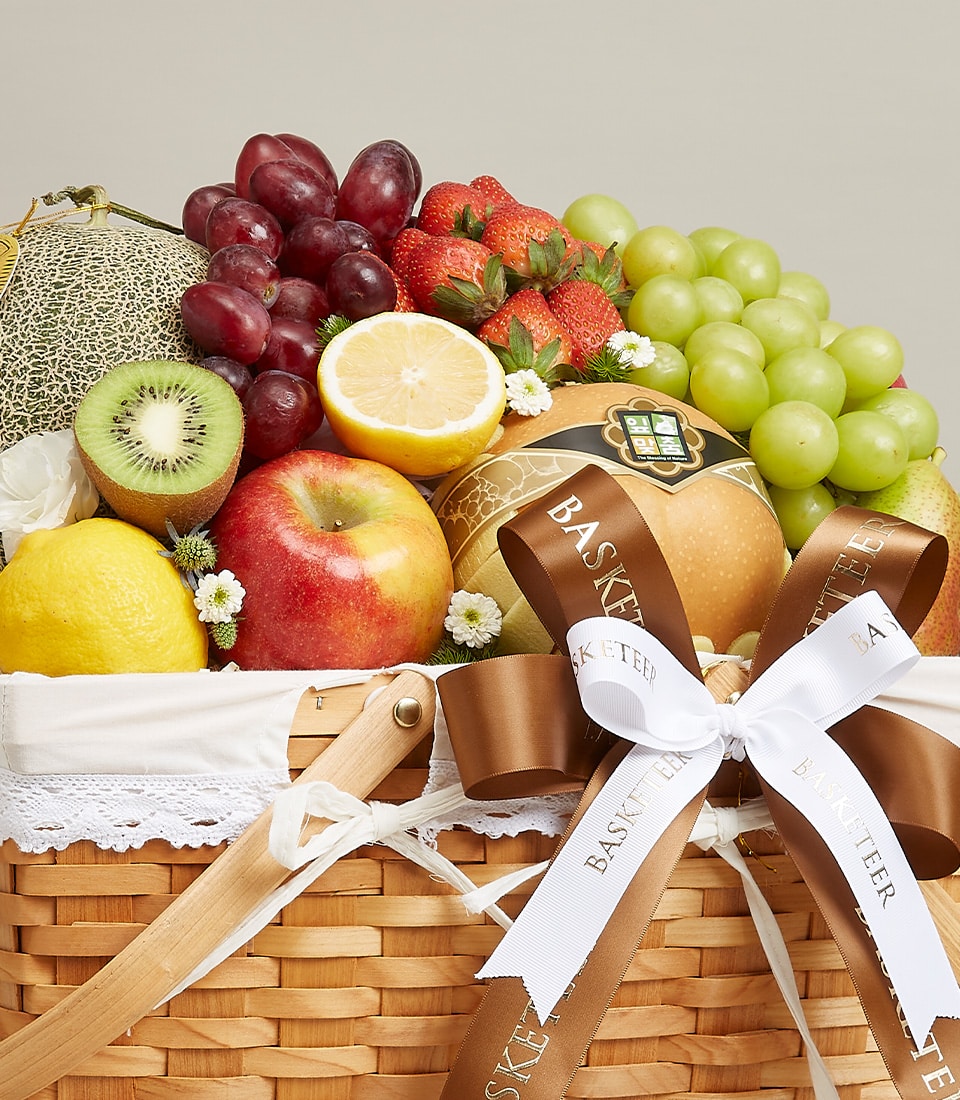 A luxurious wicker basket brimming with an exquisite assortment of handpicked fruits: luscious red and green grapes, plump strawberries, a vibrant halved orange, juicy kiwi, succulent cantaloupe slices, a crisp apple, a ripe pear, and a zesty lemon. This enchanting Artisanal Fruit Medleys Basket is elegantly adorned with delicate white lace trim and tied with a sophisticated brown ribbon bow featuring the distinguished 