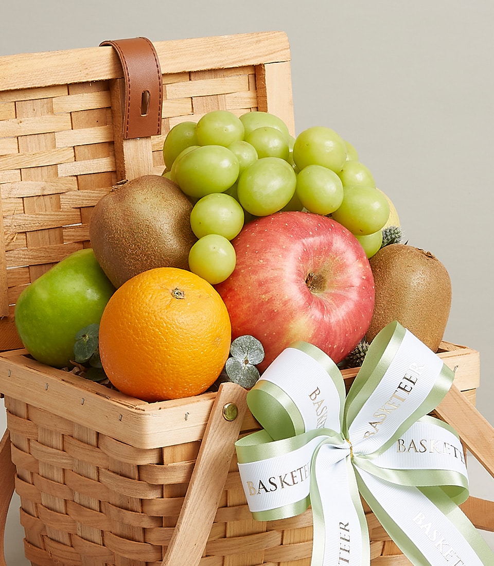 A luxurious wicker basket brimming with an array of exquisite fruits, featuring lush green grapes, a crisp red apple, a juicy green apple, a succulent kiwi, and a vibrant orange. The basket is adorned with an elegant white and green ribbon tied into a sophisticated bow and embossed with the prestigious 