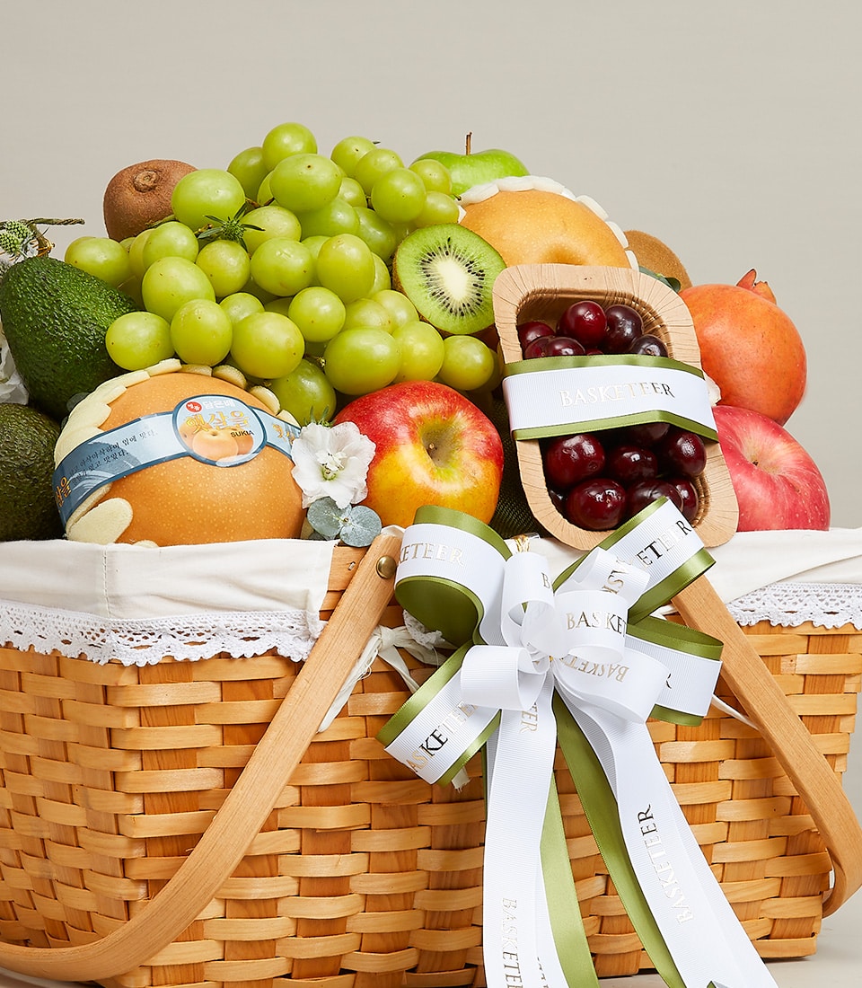 The opulent Sumptuous Fruit Bliss Basket by Basketeer is an exquisite wicker hamper brimming with a luxurious assortment of vibrant green grapes, juicy kiwis, creamy avocados, crisp pears, succulent apples, and luscious cherries. This elegantly curated collection is wrapped in sophisticated white and green ribbons and topped with a delicate decorative bow showcasing the Basketeer logo.