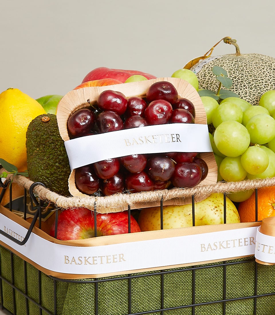 A luxurious wicker basket brimming with a tantalizing assortment of premium fruits: lush red apples, succulent green grapes, a perfectly ripe melon, and zesty lemon, alongside an exquisite avocado. In the foreground, a polished wooden tray showcases a bounty of plump, glistening red cherries adorned with an elegant white ribbon emblazoned with the prestigious Basketeer logo. This gourmet gift basket from Basketeer is the epitome of refined indulgence and is ideal for any special occasion.