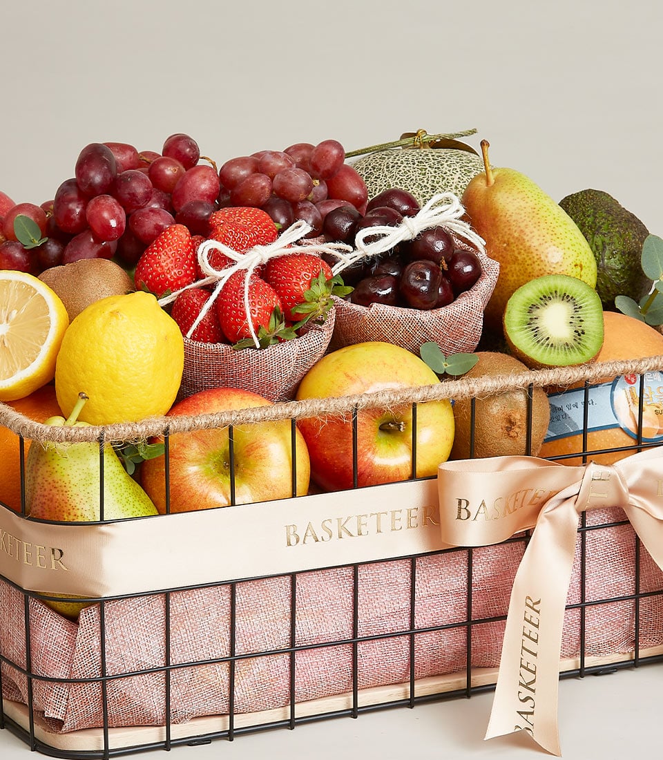 A lavish wire basket brimming with an exquisite assortment of fruits: luscious grapes, succulent pears, ripe strawberries, vibrant cherries, crisp apples, zesty lemons, exotic kiwis, and juicy melons. The basket is elegantly lined with fine fabric and adorned with a cream-colored ribbon bearing the prestigious 