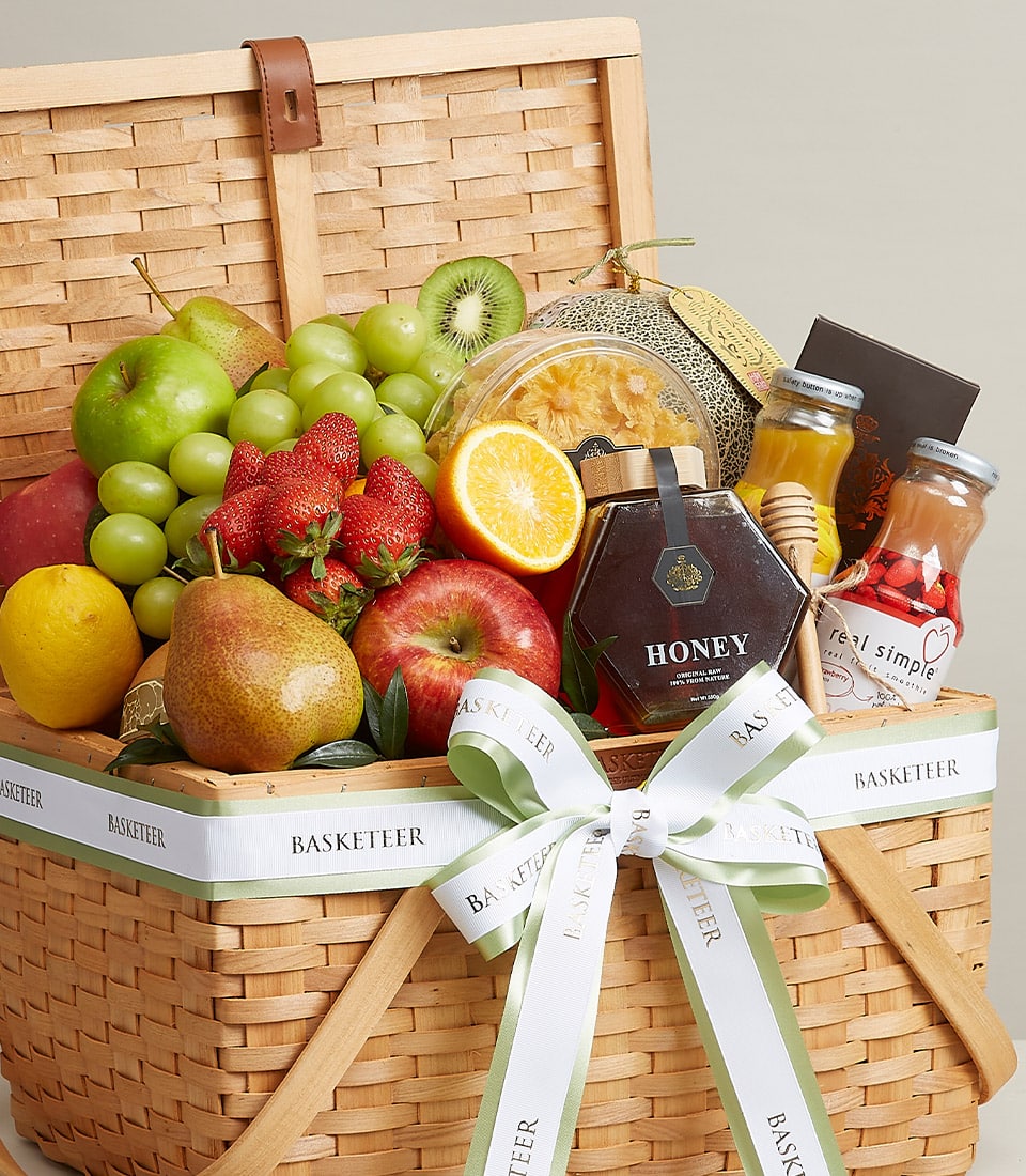 A luxurious wicker basket brimming with an exquisite selection of fresh fruits—crisp apples, succulent pears, luscious grapes, juicy strawberries, and a perfectly sliced orange. Nestled within are jars of golden honey and gourmet preserves, accompanied by two bottles of refreshing juice. The elegant ensemble is adorned with a sophisticated green and white ribbon that features the distinguished Basketeer logo, elevating the scene to one of pure indulgence.