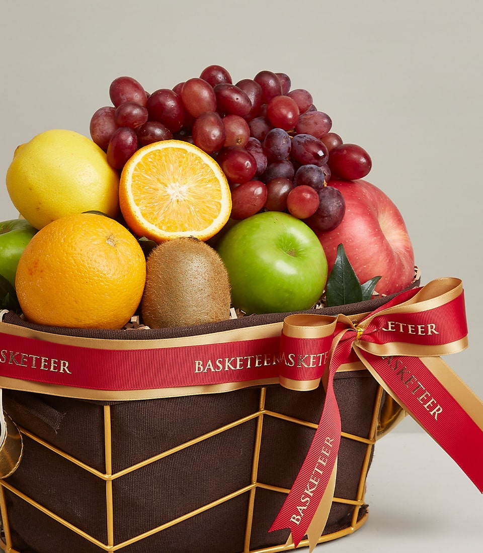 A luxurious wicker basket overflowing with a delightful assortment of premium fruits, including plump red grapes, a juicy orange half, crisp green and red apples, a succulent kiwi, and zesty lemons. The basket is elegantly adorned with a sophisticated red and gold ribbon proudly bearing the 