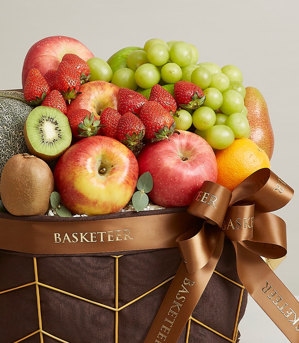 A beautifully curated Mixed Fruit Basket from Basketeer that exudes luxury and charm. The basket features an exquisite assortment of the freshest apples, luscious strawberries, vibrant green grapes, succulent kiwi, a fragrant melon, and a zesty orange. This gourmet selection is elegantly presented with a rich brown ribbon adorned with the BASKETEER logo, adding a sophisticated touch. Set against a serene and neutral backdrop, this opulent offering invites indulgence and sophistication.