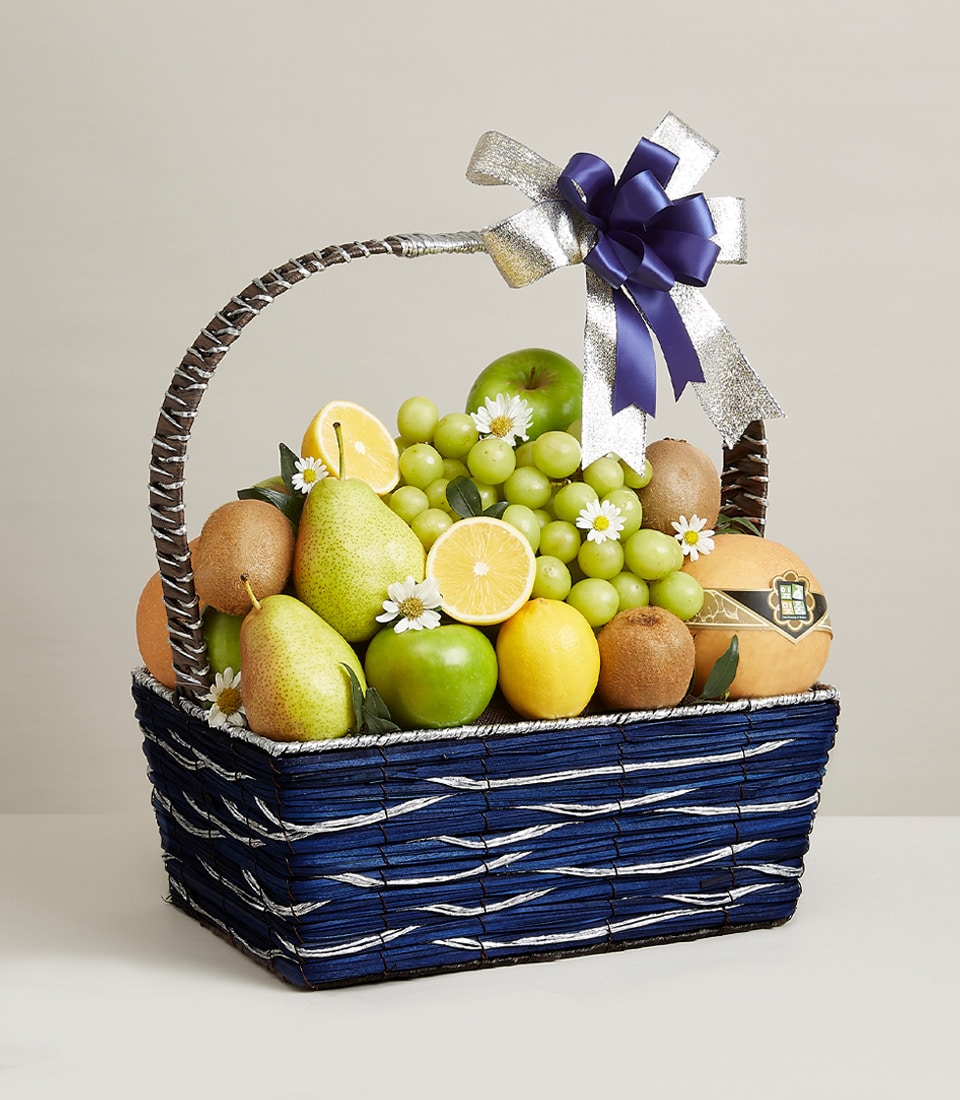 Presenting The Royal Fruit Delights Basket by Basketeer: a sophisticated blue woven basket brimming with an exquisite selection of premium fruits—lush grapes, succulent pears, zesty lemons, crisp apples, and exotic kiwis. Elegantly adorned with a shimmering silver and regal purple bow, this luxurious gift ensemble is further enhanced by delicate white and yellow blossoms nestled among the fruits. The opulence is unmistakable as the Basketeer logo graces the accompanying ribbon and box, making it a truly inviting indulgence perfect for any refined occasion.