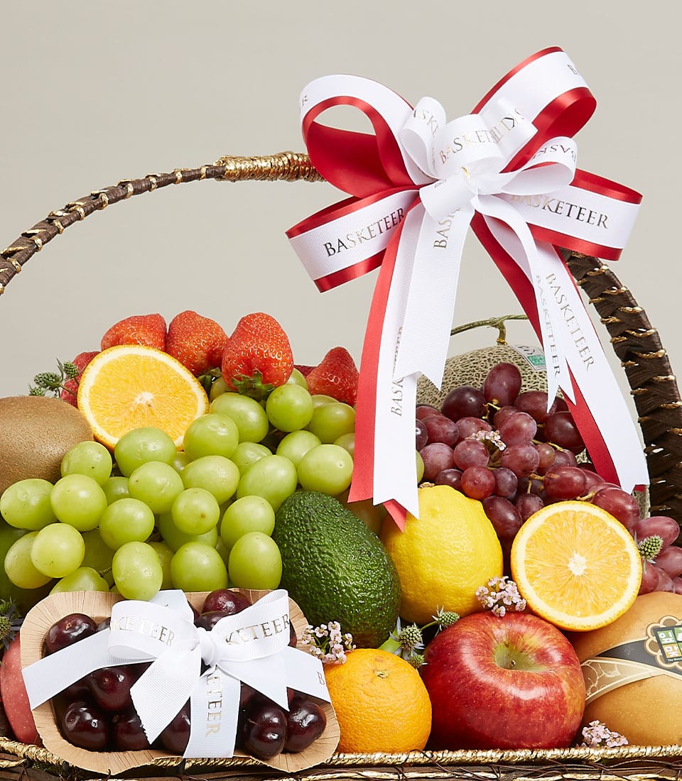 A sumptuous wicker basket, meticulously arranged with the freshest assortment of green and red grapes, a ripe avocado, a crisp apple, a zesty lemon, succulent kiwis, juicy strawberries, vibrant orange slices, and plump cherries. This luxurious fruit basket is elegantly decorated with a sophisticated white and red ribbon proudly displaying the 