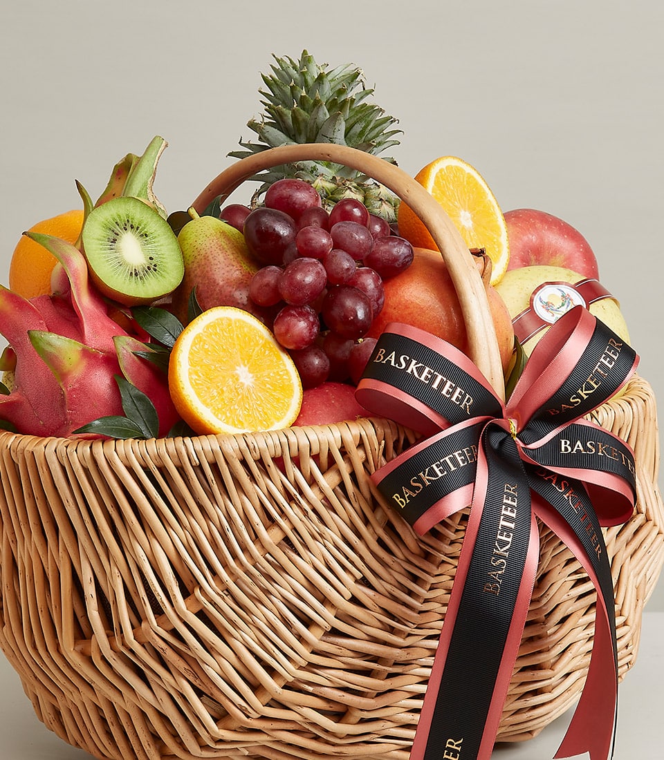 A luxurious wicker basket, adorned with an elegant BASKETEER ribbon tied into a graceful bow, brims with an exquisite selection of orchard-fresh fruits. Among the vibrant bounty are exotic dragon fruit, succulent oranges, luscious kiwifruit, plump grapes, and a majestic pineapple. The scene is set against a sophisticated light grey backdrop, inviting you to indulge in the premium freshness and quality curated by Basketeer.