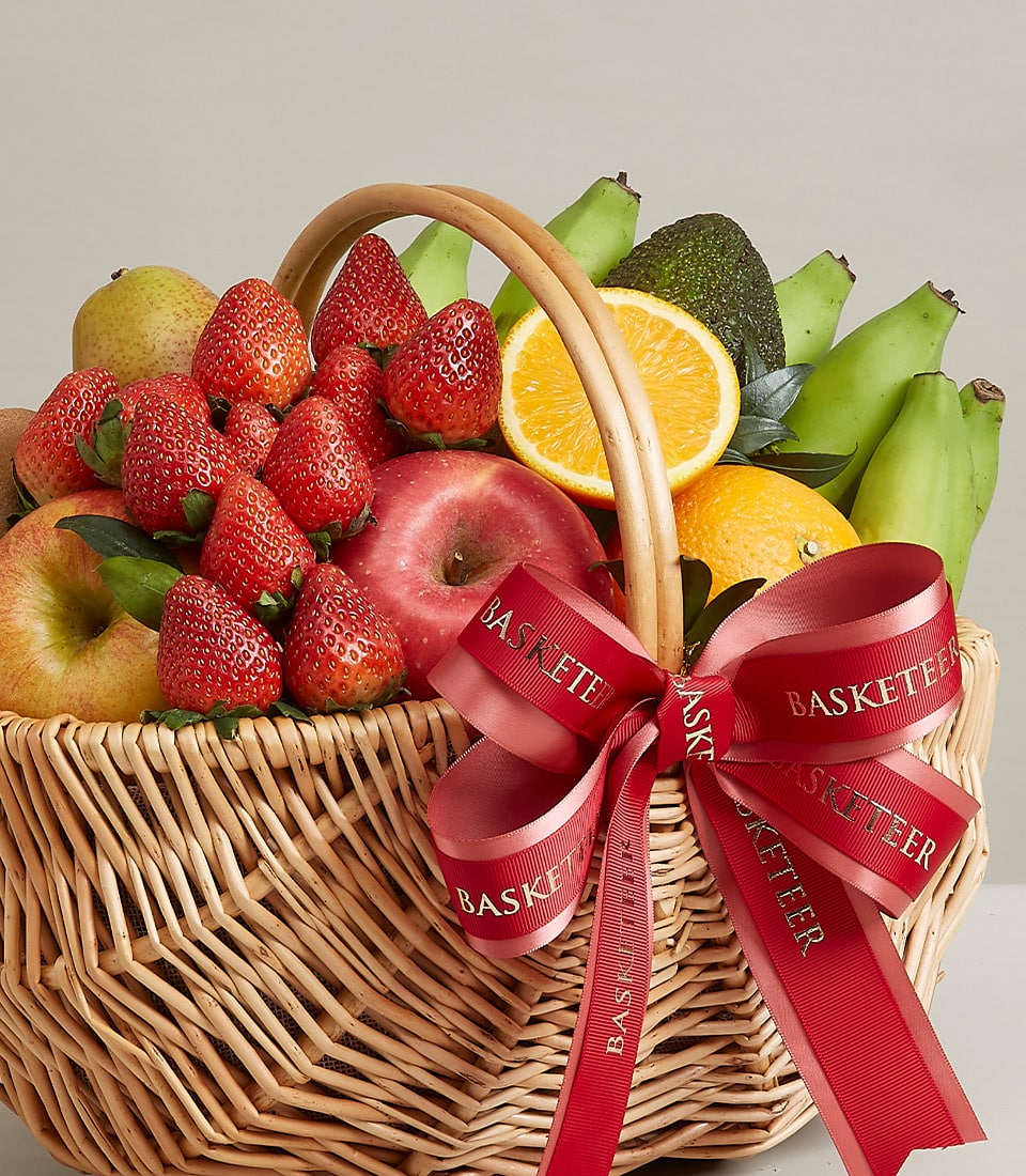 A lavish wicker basket brimming with a vibrant array of fresh, hand-picked fruits, featuring succulent strawberries, crisp apples, juicy pears, ripe bananas, a perfectly halved orange, and creamy avocados. This beautifully elevated arrangement is elegantly adorned with a sophisticated red ribbon showcasing the distinguished 