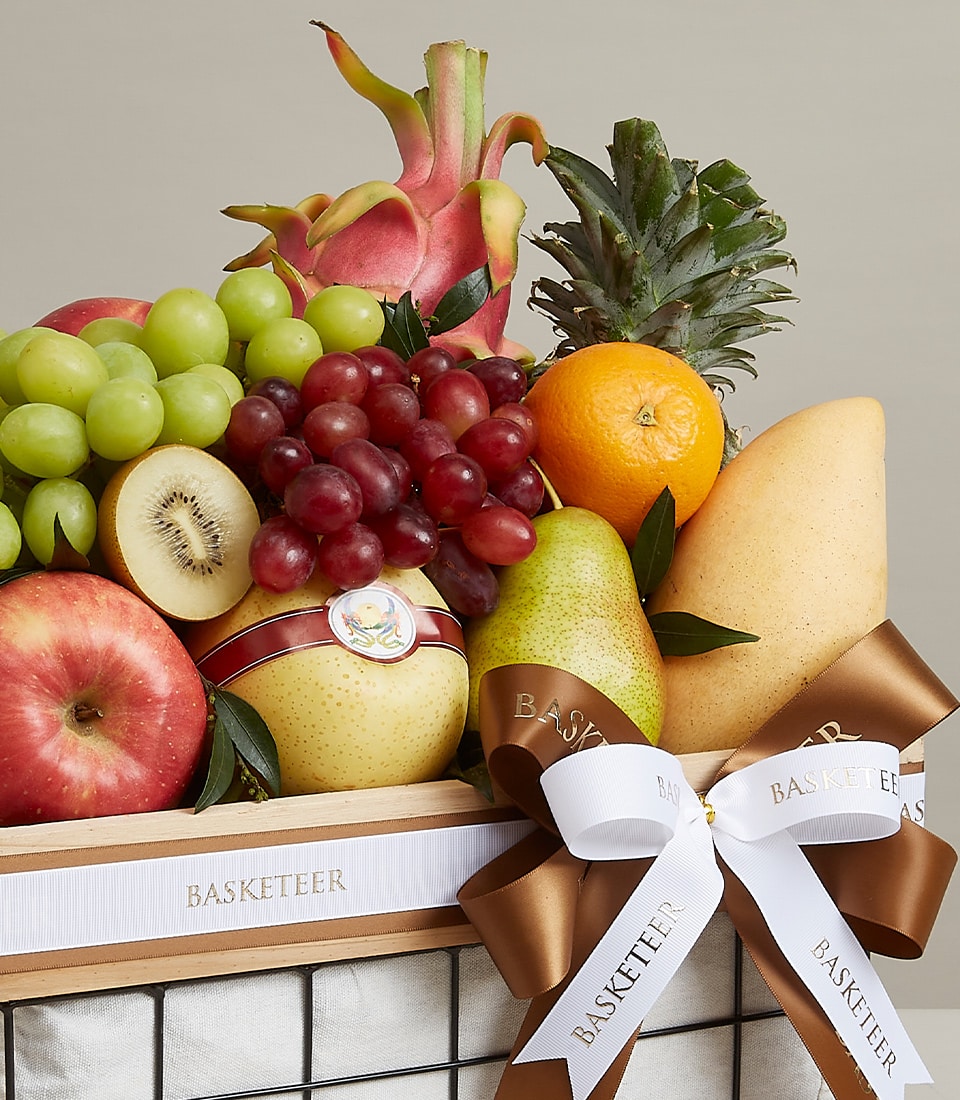 An opulent fruit gift basket by Basketeer, elegantly decorated with a brown and white ribbon bearing the brand's signature logo. This meticulously curated selection bursts with vibrant, fresh fruits: lush green and red grapes, an exotic dragonfruit, a regal pineapple, a zesty orange, a crisp apple, a succulent pear, and a ripe melon. The luxurious presentation exudes sophistication and invites delight.