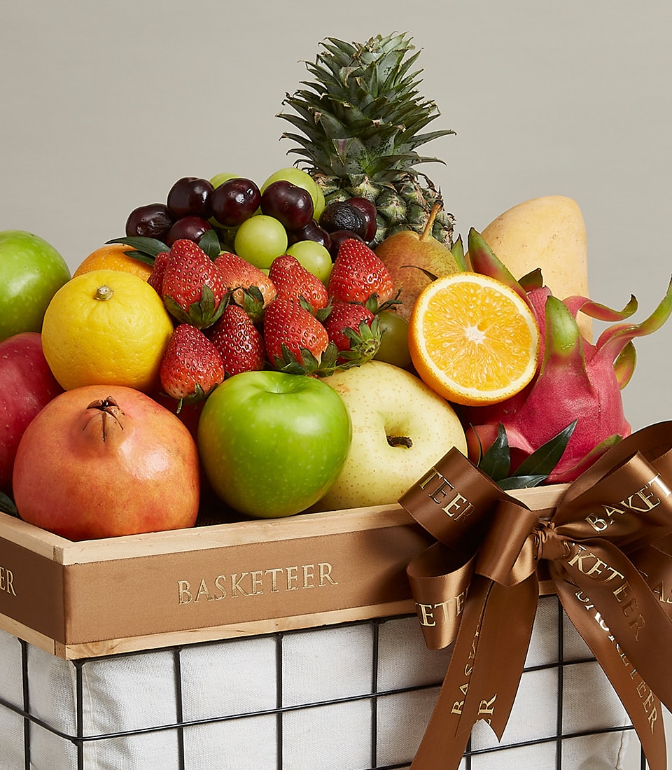 A luxurious fruit ensemble basket brimming with an exquisite selection of fresh fruits, including succulent strawberries, crisp apples, zesty oranges, plump grapes, exotic dragon fruit, ripe pineapple, tangy lemon, and vibrant pomegranate. The elegant wooden crate is adorned with a sophisticated brown ribbon and rests gracefully atop a pristine white cloth. The handle proudly features the distinguished 