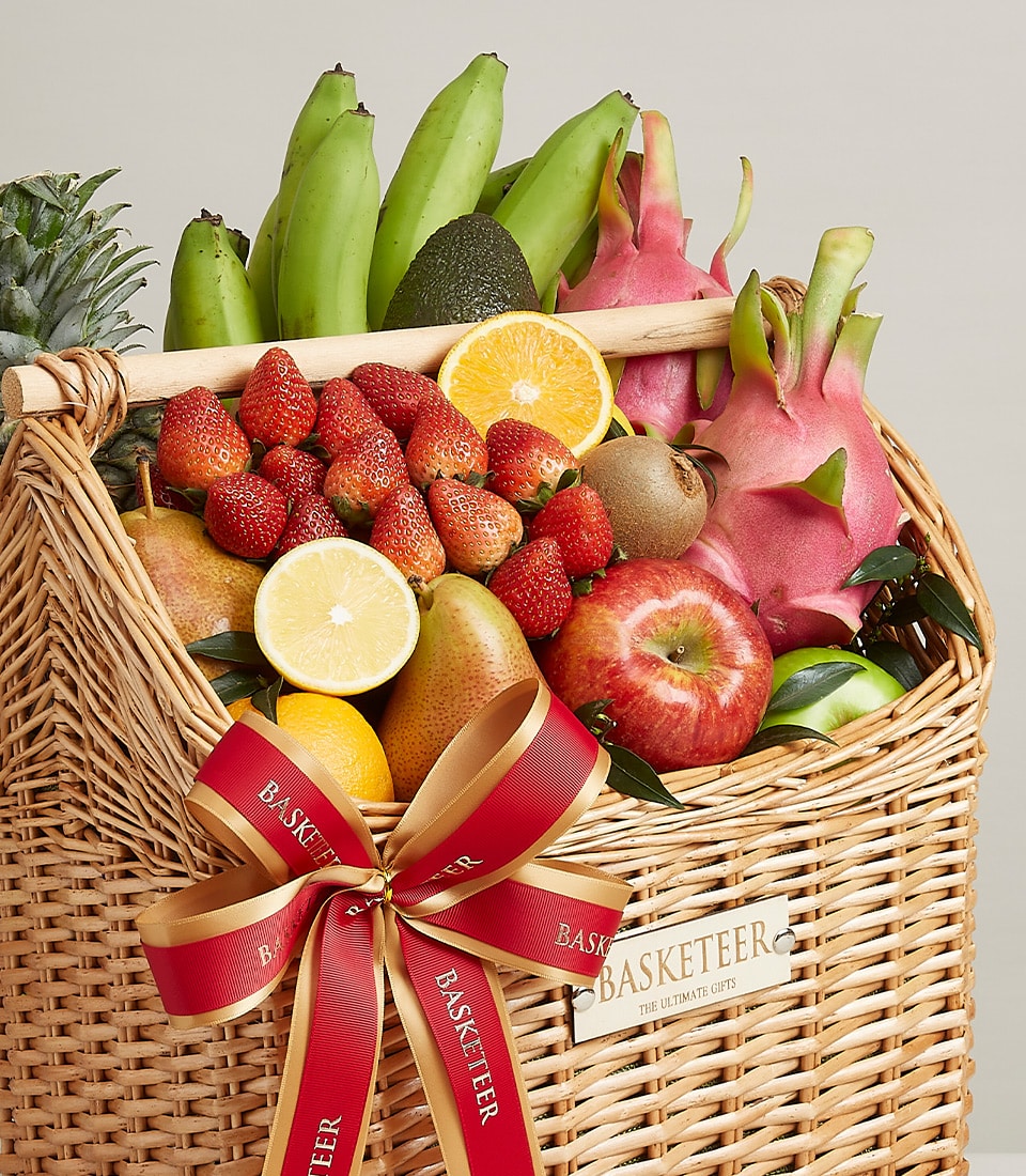 An exquisite woven basket overflowing with a vibrant assortment of premium fresh fruits, including succulent strawberries, perfectly ripe bananas, luscious pineapple, zesty oranges, crisp apples, exotic dragonfruit, and creamy avocados. The basket is elegantly adorned with a luxurious red and gold ribbon and a tag that proudly displays 