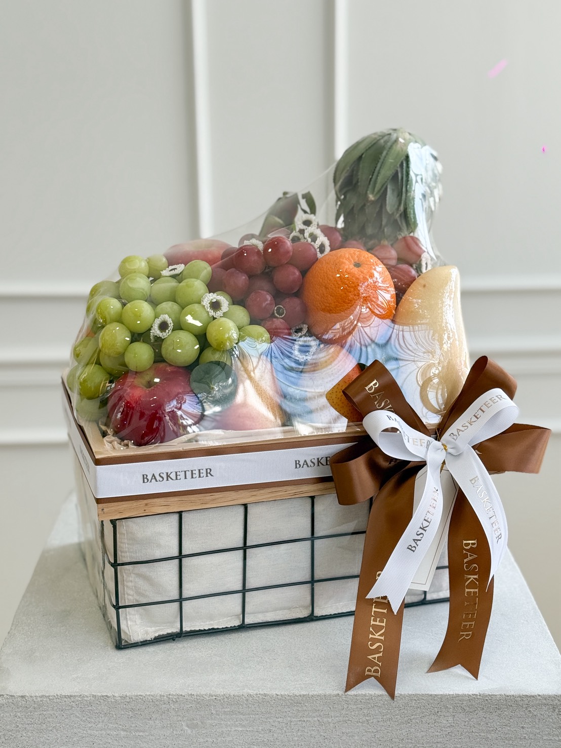 Luxury fresh fruit basket with grapes, oranges, pears, apples, bananas, and tropical fruits, decorated with brown and white satin ribbon