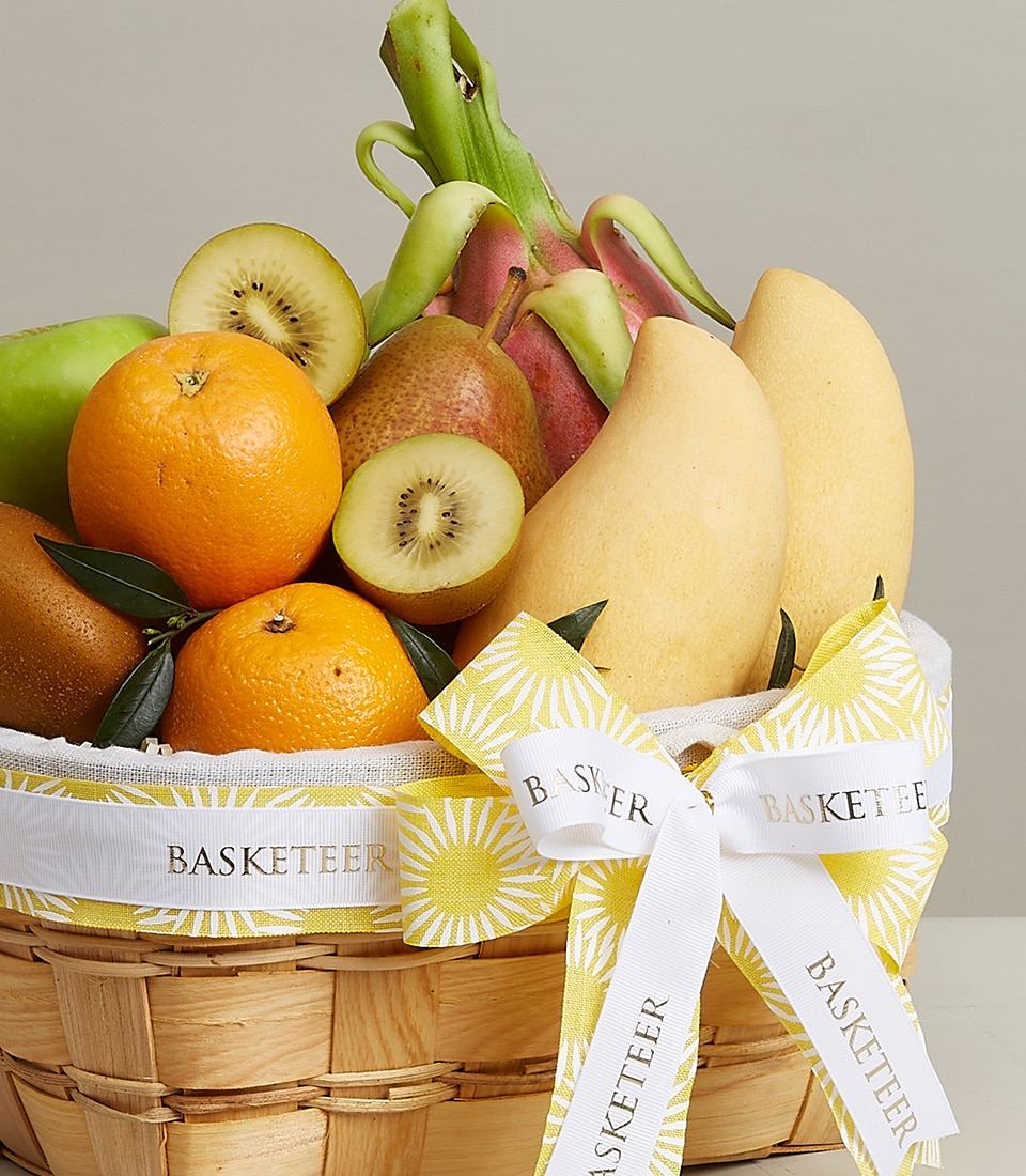 A lavish gourmet fruit basket overflowing with a curated selection of premium fruits, including vibrant oranges, luscious kiwis, elegant pears, an exotic dragon fruit, and two golden-hued mangoes. The sophisticated wicker basket is tastefully adorned with a luxurious white and yellow ribbon showcasing the prestigious 