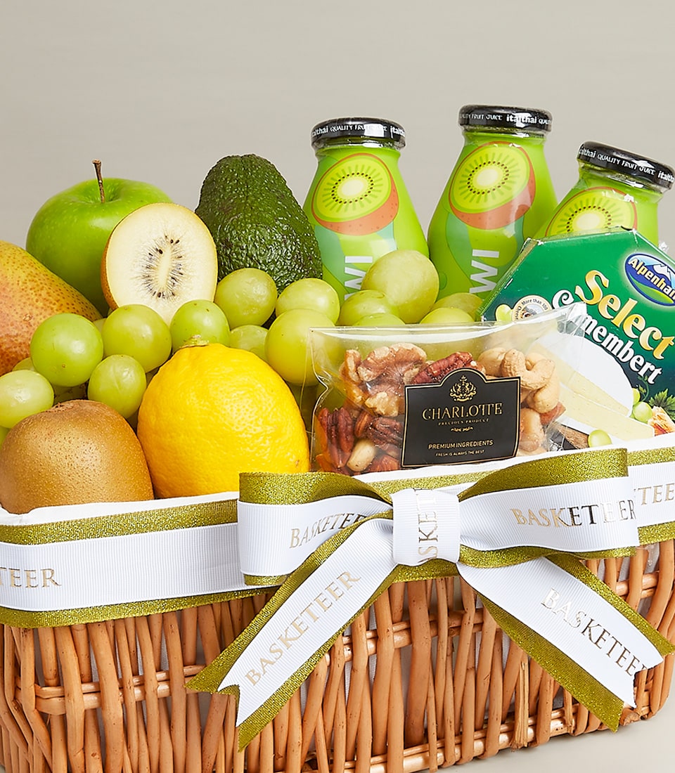 A luxurious wicker basket overflowing with nature’s finest offerings, including succulent green grapes, a vibrant lemon, a crisp green apple, a perfectly ripe avocado, a kiwi elegantly halved to reveal its emerald core, and an opulent pear. Tucked among this bounty are premium bottles of kiwi drink and an artisanal bag of mixed nuts. The arrangement is exquisitely adorned with a sophisticated white and green ribbon featuring the Basketeer logo and the inscription 