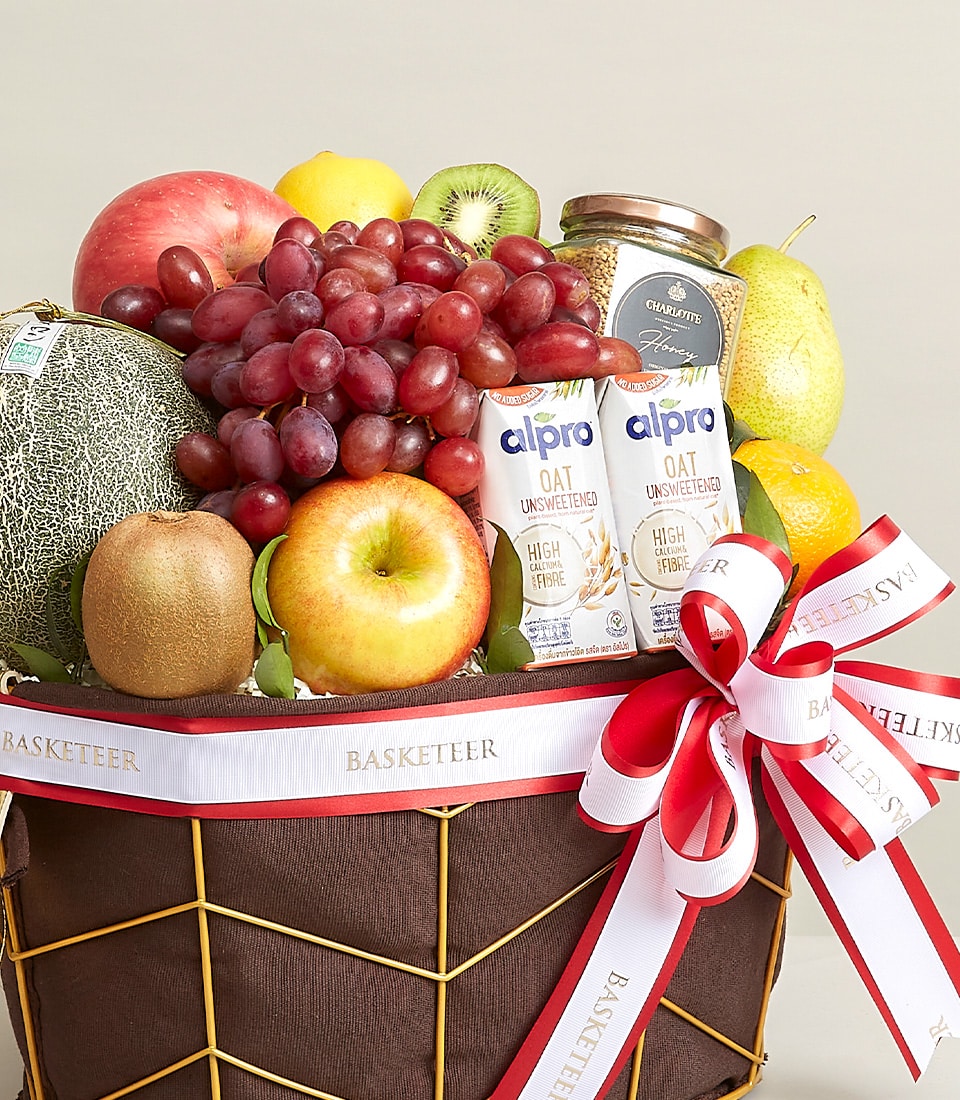 A lavishly curated fruit basket overflowing with an exquisite selection of apples, melons, grapes, pears, and kiwi. Two cartons of Alpro oat milk elegantly complement the bounty. The entire ensemble is adorned with a luxurious red and white ribbon featuring the prestigious 