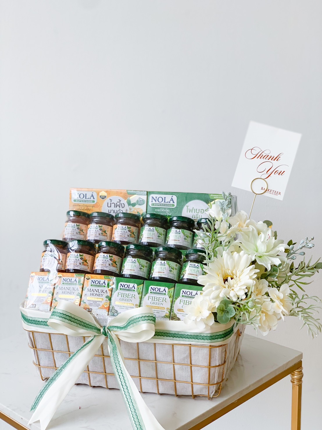 A gift basket contains various jars of NOLA-branded food products, with packaging labels in what appears to be Thai. The basket is adorned with a green and white ribbon and a bouquet of white flowers. A 
