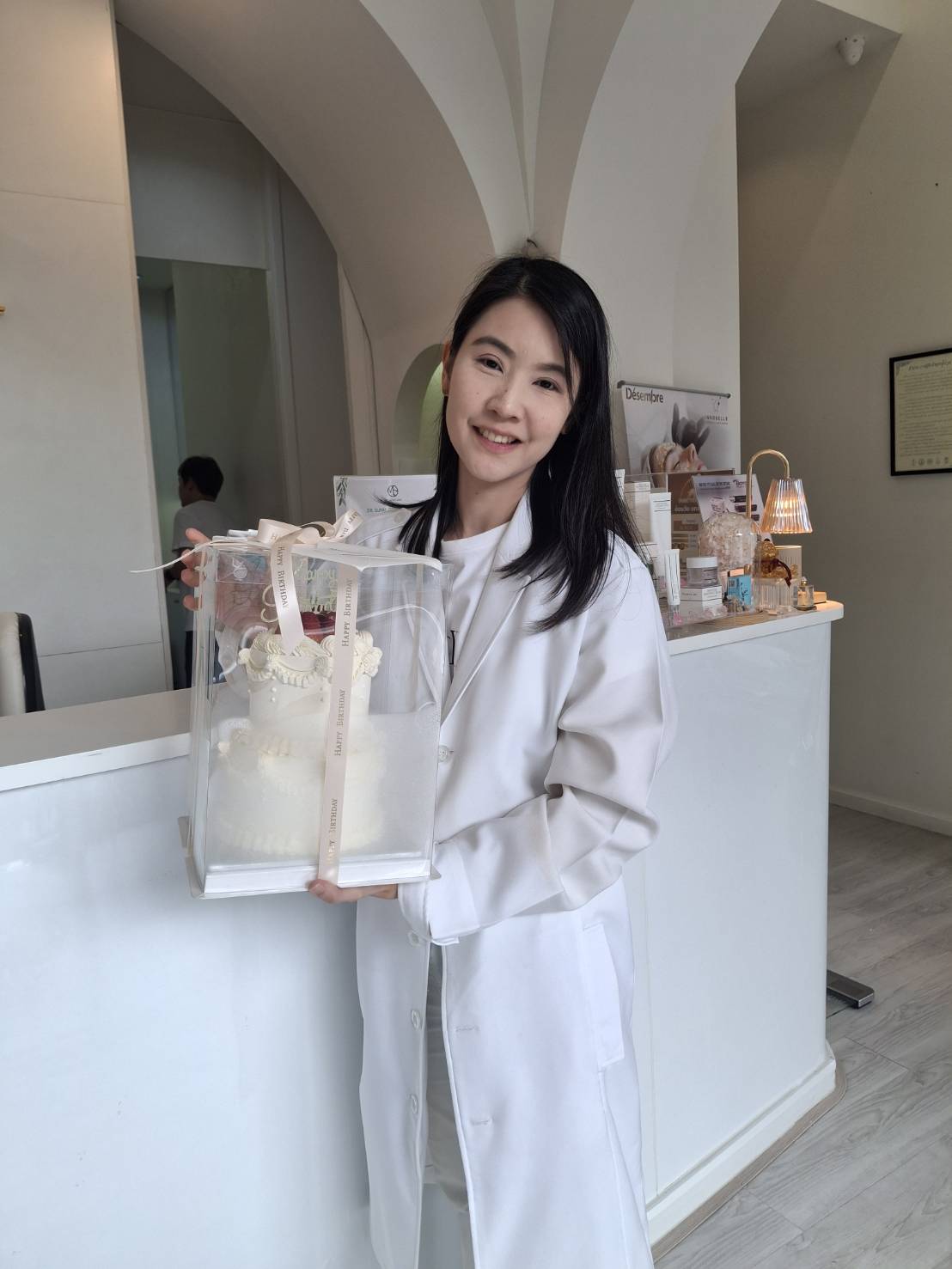 A woman in a white coat is standing inside a brightly lit room, holding a boxed, intricately decorated cake perfect for a princess. She is smiling and standing in front of a white reception counter with various products and decor items.