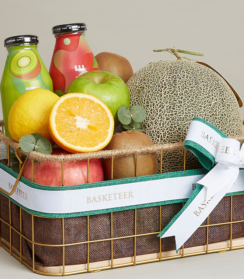 A luxurious and elegantly arranged decorative basket brimming with a vibrant assortment of fresh, succulent fruits, including a whole ripe cantaloupe, a crisp green apple, half a juicy orange, a luscious red apple, and an exotic kiwi. Nestled among this delightful bounty are two gourmet bottles of premium fruit juice. The basket is tastefully adorned with an exquisite green and white ribbon proudly showcasing the 