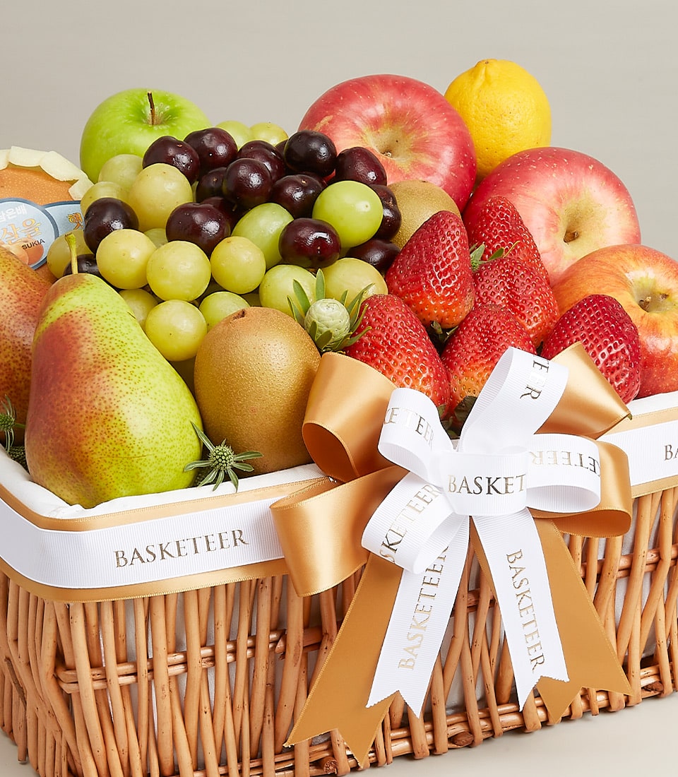 An exquisite, premium fresh fruit basket by Basketeer, brimming with a selection of lush green and red apples, plump grapes, succulent strawberries, crisp pears, a vibrant kiwi, a zesty lemon, and a juicy orange. This luxurious wicker basket is elegantly adorned with a sophisticated white and gold ribbon proudly displaying the 