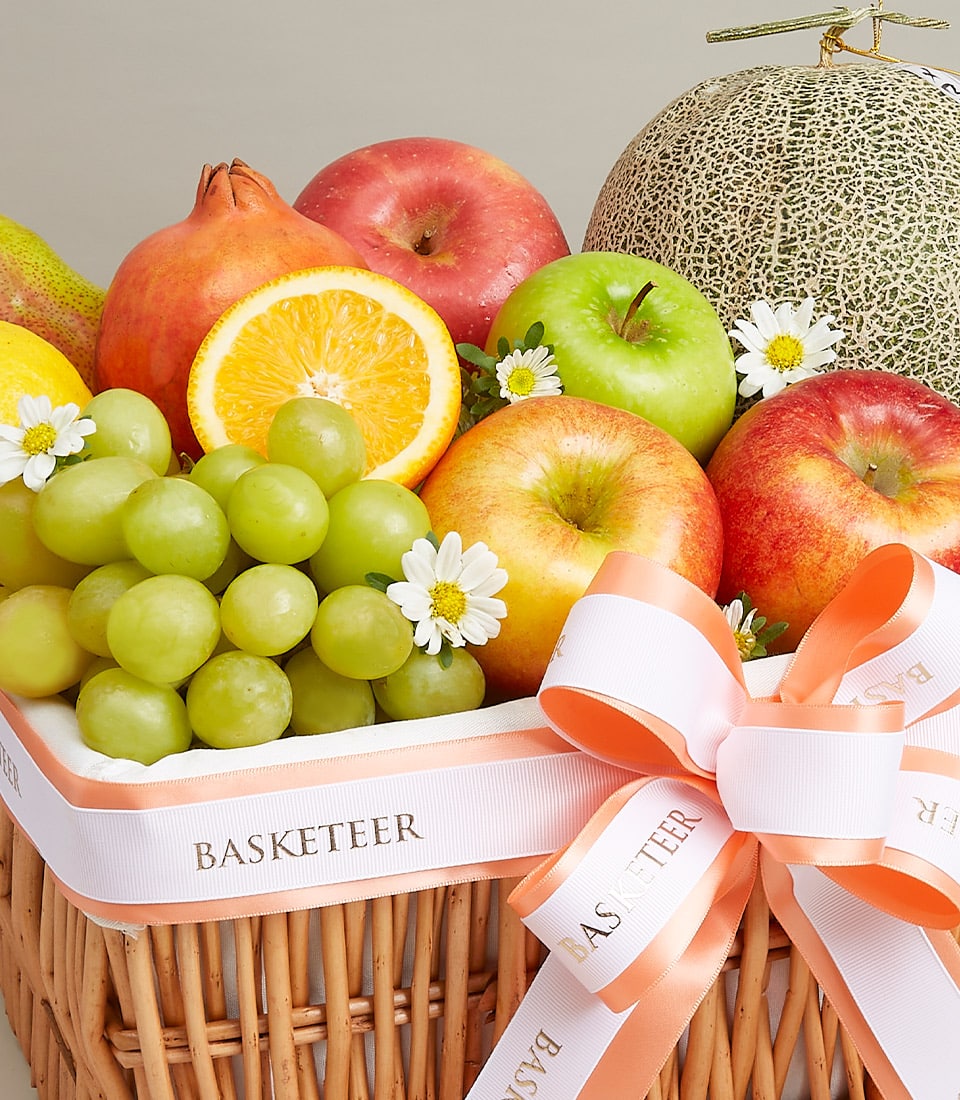 A sumptuous Fresh Fruits Gift Basket, impeccably curated with a delightful assortment of premium fruits: luscious green grapes, crisp apples, juicy pears, a ripe cantaloupe, and a tantalizing slice of orange. This exquisite bounty is elegantly presented in a woven basket, adorned with delicate white blossoms and finished with an opulent peach-colored ribbon proudly displaying the prestigious 