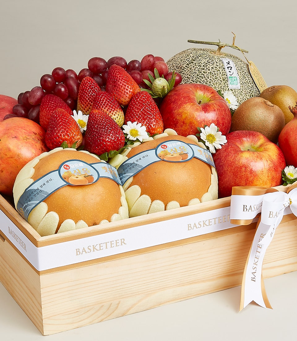 A luxurious wooden gift box adorned with the elegant Basketeer logo, overflowing with an exquisite assortment of premium fruits: luscious grapes, juicy strawberries, crisp apples, succulent Asian pears, and a perfectly ripened melon. The Mixed Fruit Joy Gift Box is meticulously decorated with delicate white flowers and a refined ribbon that adds a touch of sophistication.