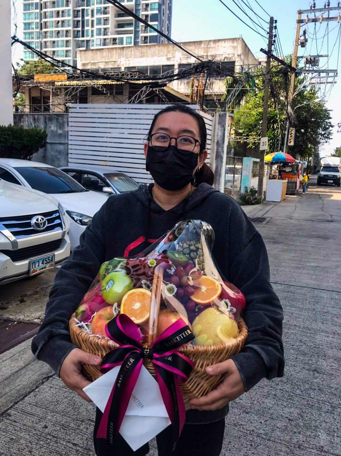 A person standing outdoors holding an Orchard Fresh Fruits Hamper filled with various fruits wrapped in clear plastic and tied with pink and black ribbons. The person is wearing glasses, a black face mask, and a dark hoodie. Buildings and parked cars are visible in the background.