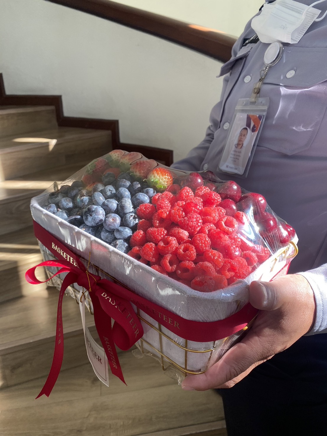 A luxurious golden wire basket filled with fresh raspberries, blueberries, and strawberries, wrapped with a red ribbon and Basketeer tag.