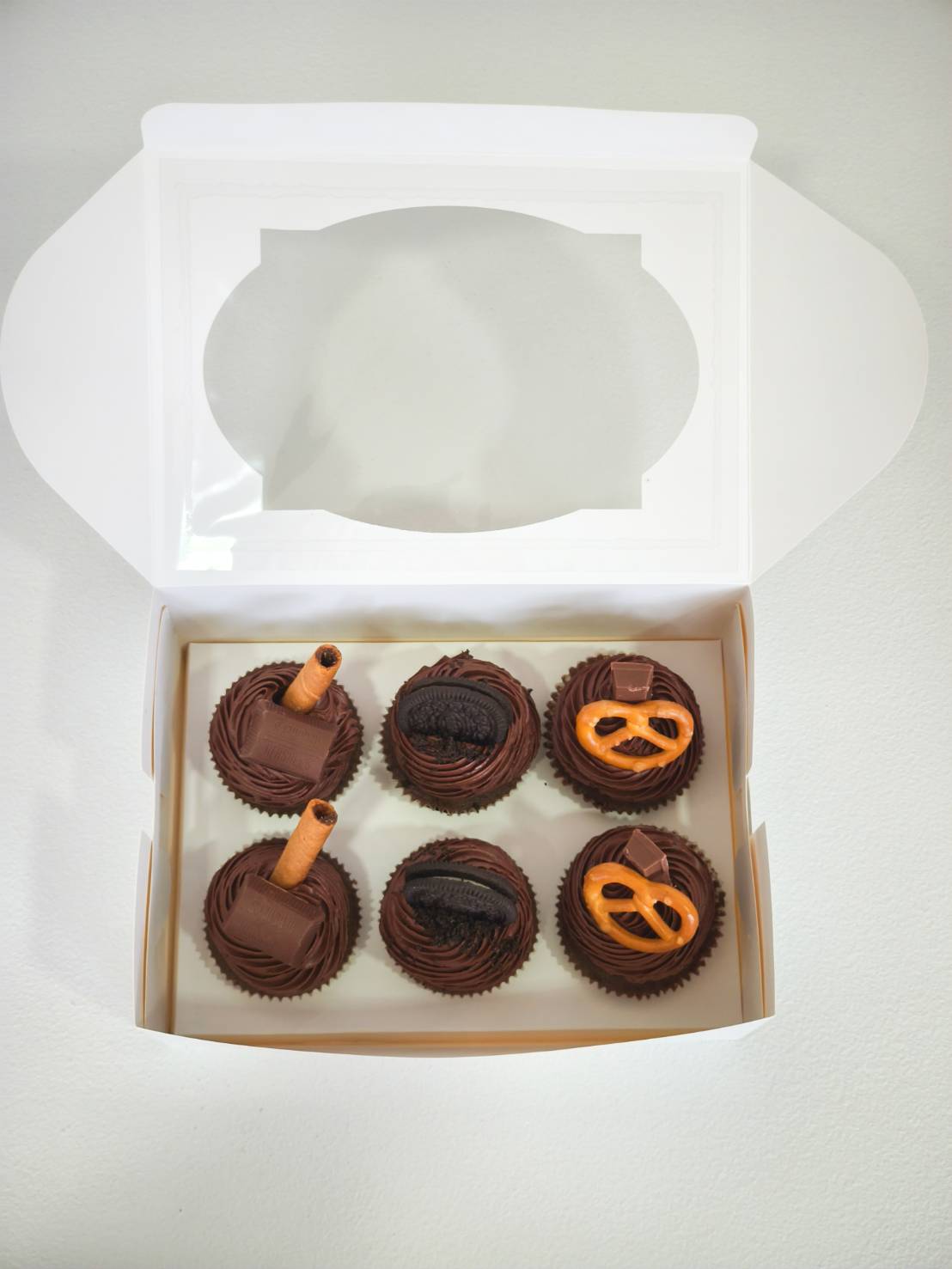 Box of six chocolate cupcakes topped with Oreo, pretzel, chocolate bars, and wafer rolls in white windowed packaging.
