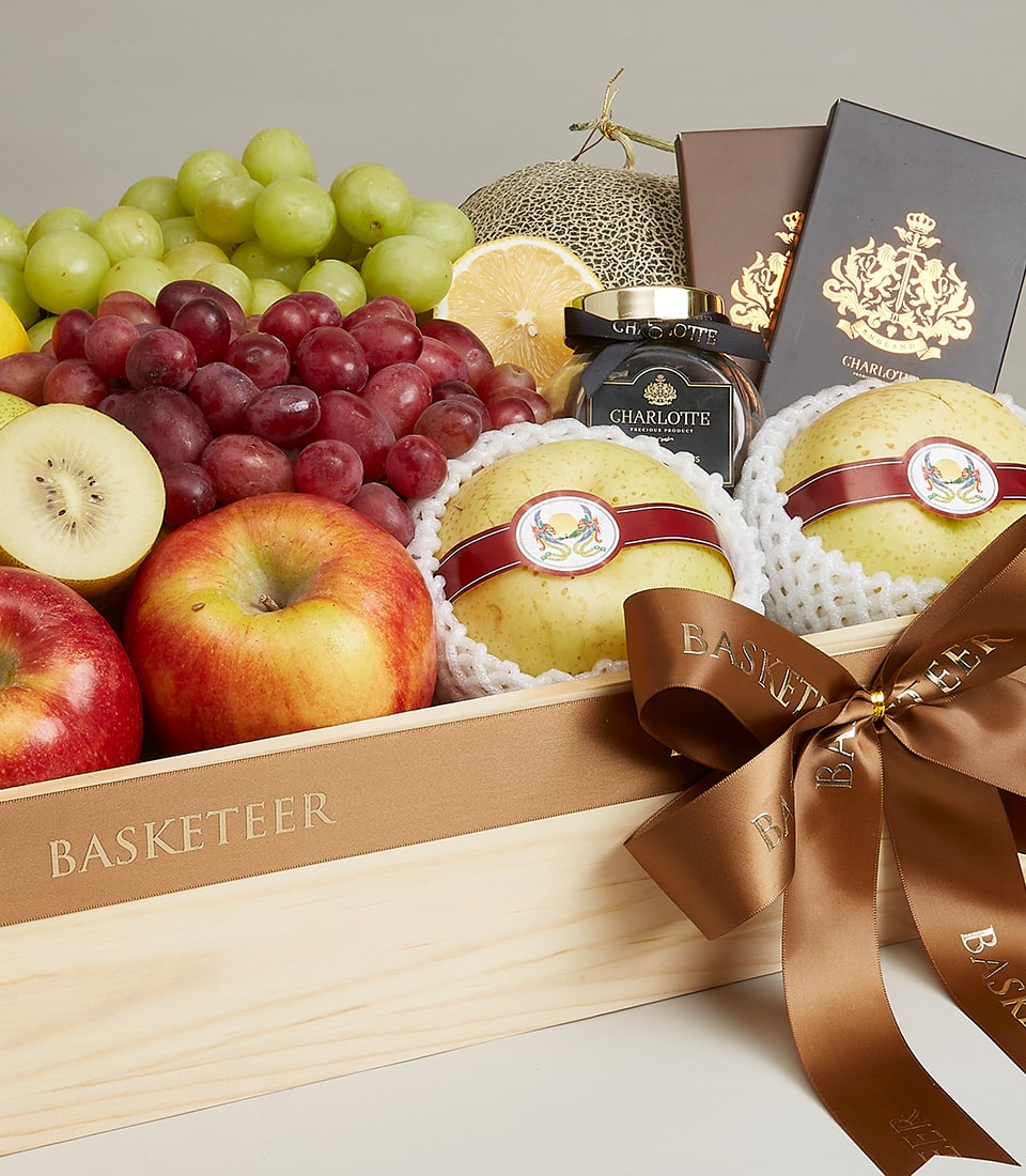 A lavish wooden gift box by Basketeer steals the spotlight, adorned with an elegant brown ribbon and exuding sophistication. Nestled within the box is a curated selection of premium fruits—succulent green and red grapes, crisp apples, exotic kiwi, and a tantalizing melon. Accompanying these fresh delights are artisanal jars of fruit preserves and two indulgent chocolate bars, offering a perfect balance of natural sweetness and decadent luxury. The Basketeer logo subtly graces the scene, promising nothing but the finest quality for its discerning recipients.