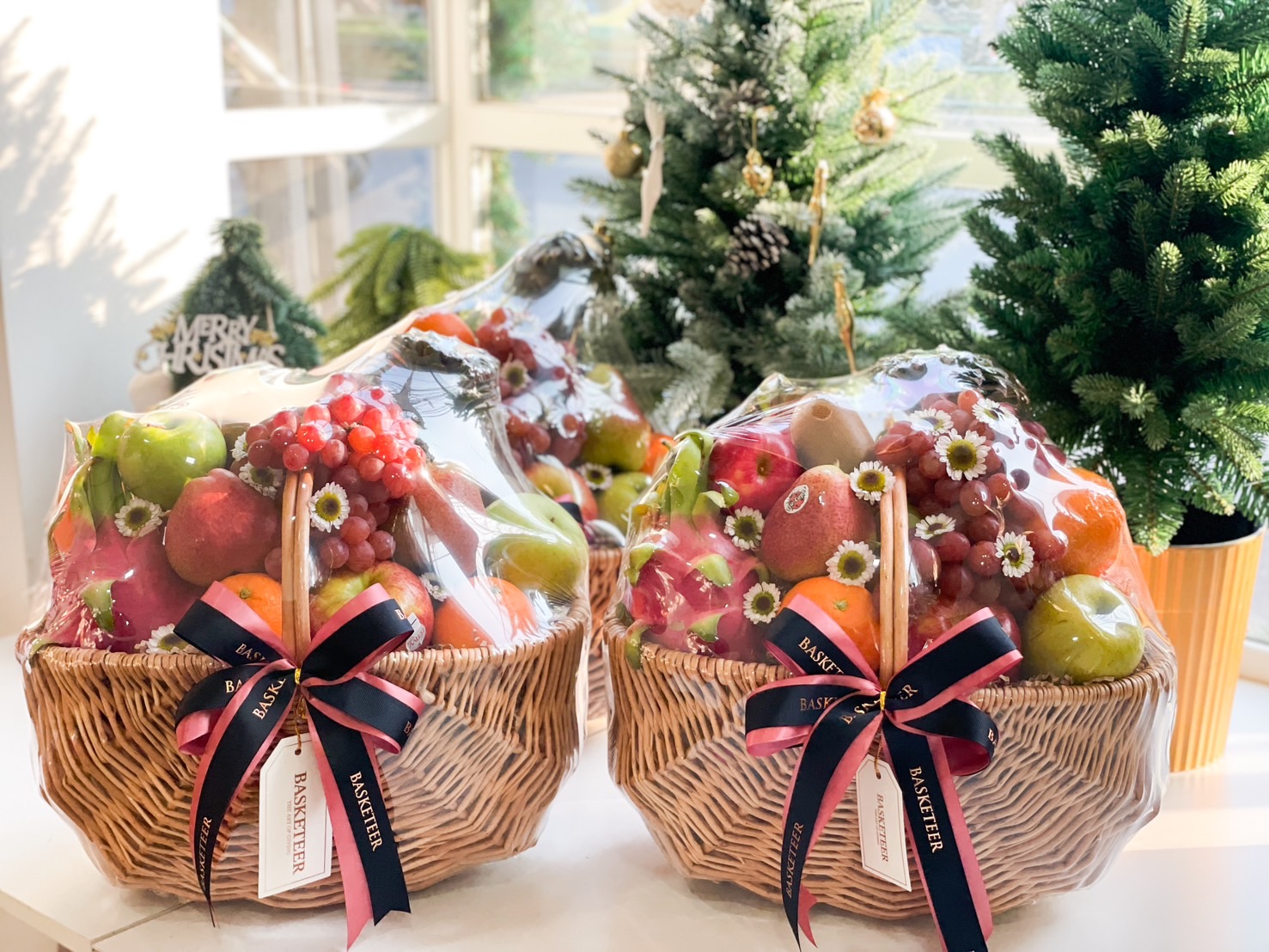 Two beautifully arranged fruit baskets filled with apples, grapes, oranges, and pears, wrapped in clear film and tied with navy and pink ribbons, set against a festive background.
